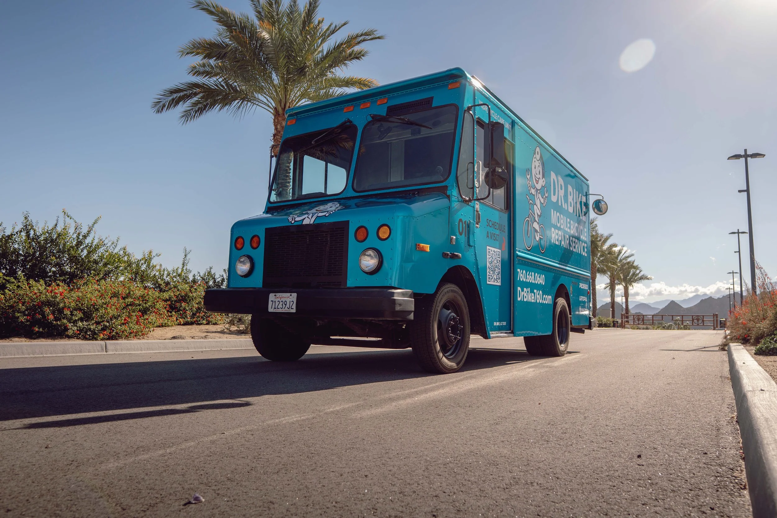 A blue Dr. Bike mobile bicycle repair truck parked on a paved path with palm trees and mountain scenery in the background.