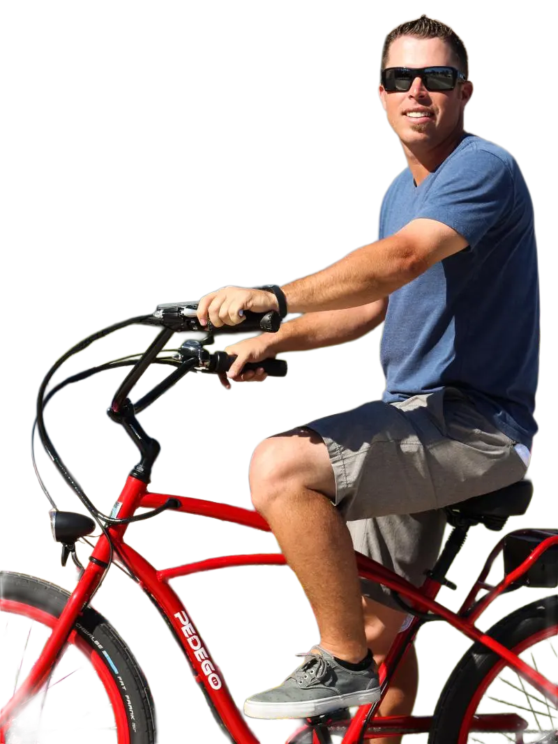 A man wearing sunglasses, a blue t-shirt, and gray shorts riding a red Pedego electric bicycle outdoors.