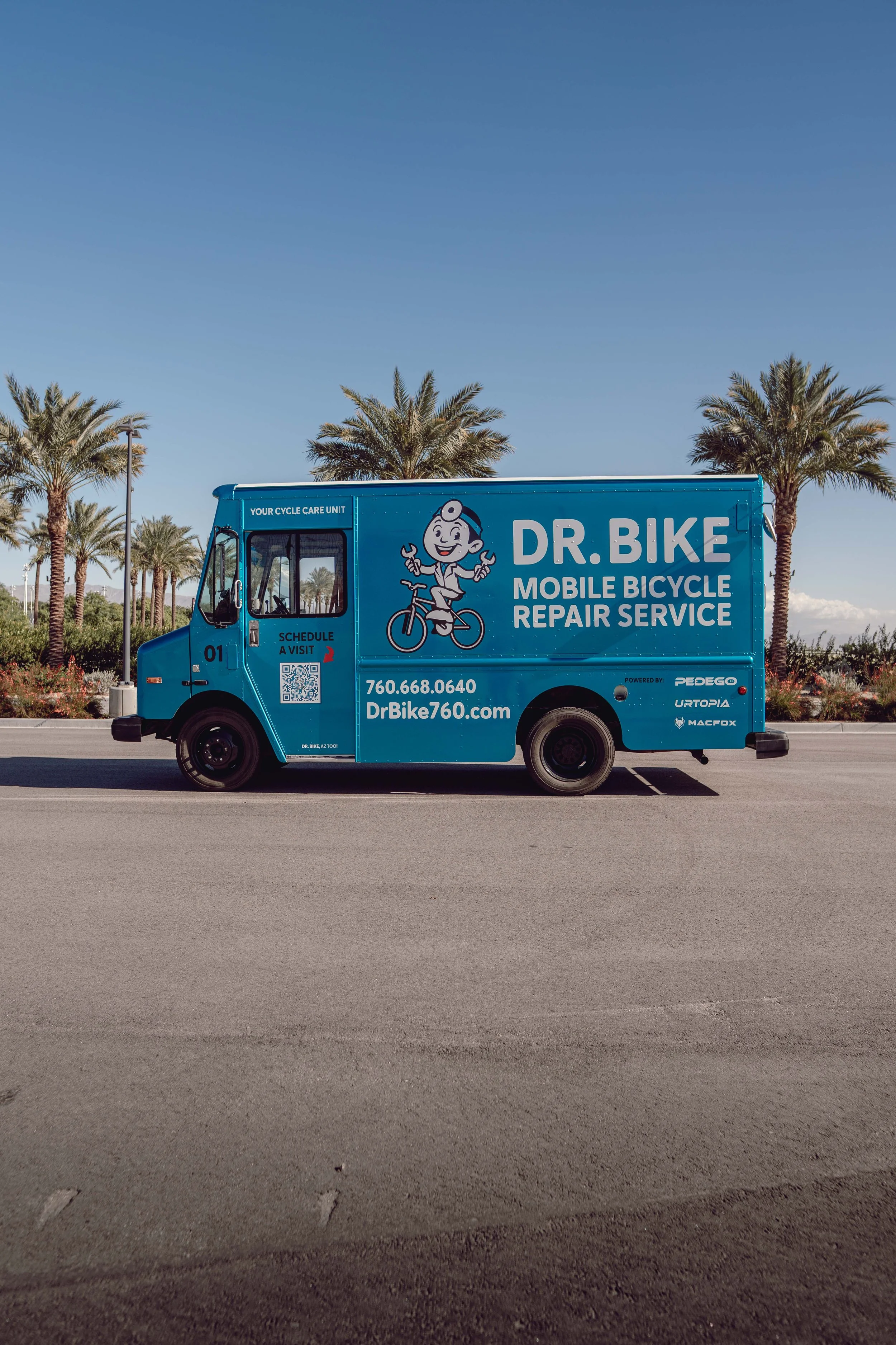 A blue mobile bicycle repair service truck parked on an empty street with palm trees and a clear blue sky in the background.