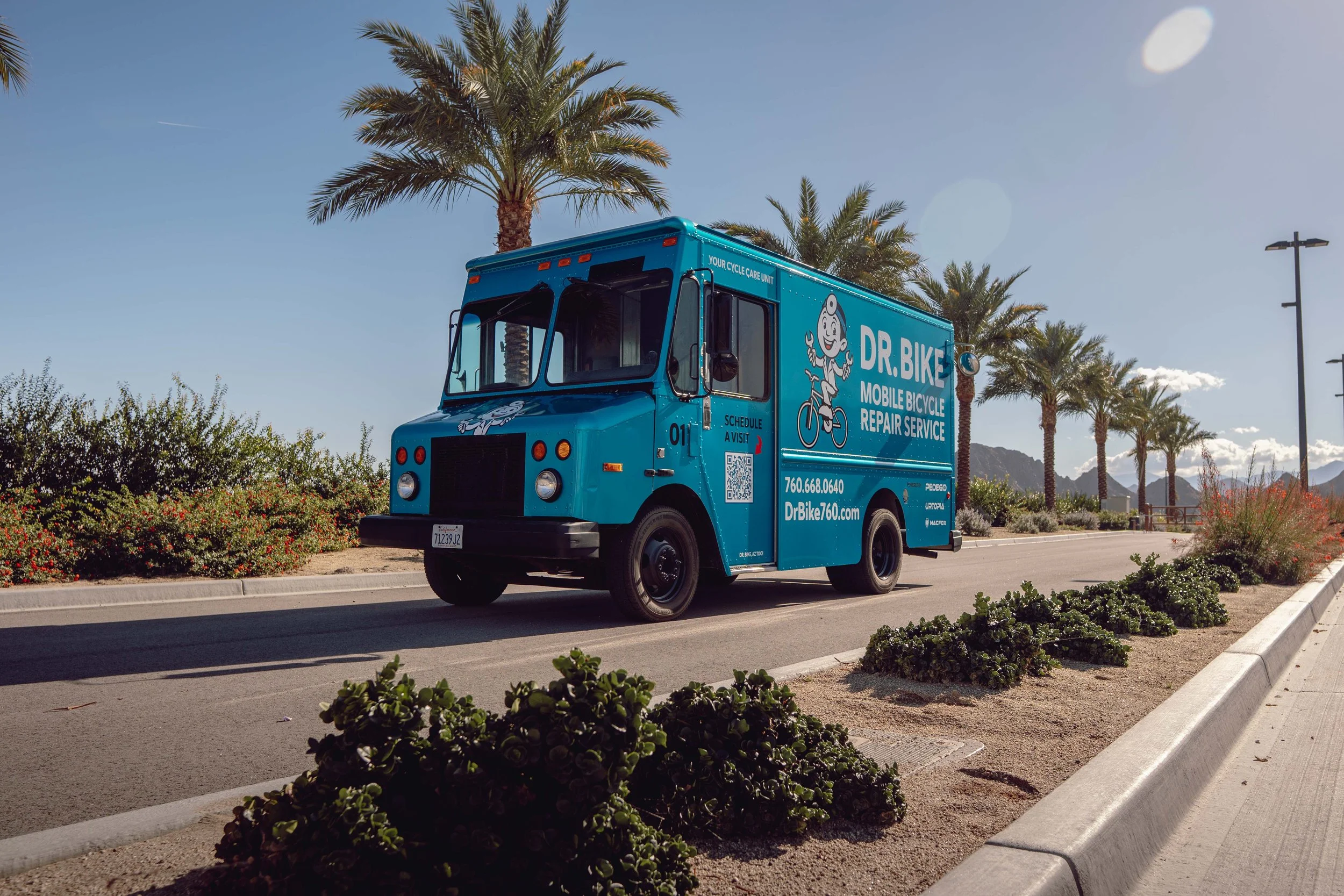 A bright blue mobile bicycle repair service truck labeled 'DR. BIKE' parked on a street with palm trees and mountains in the background.