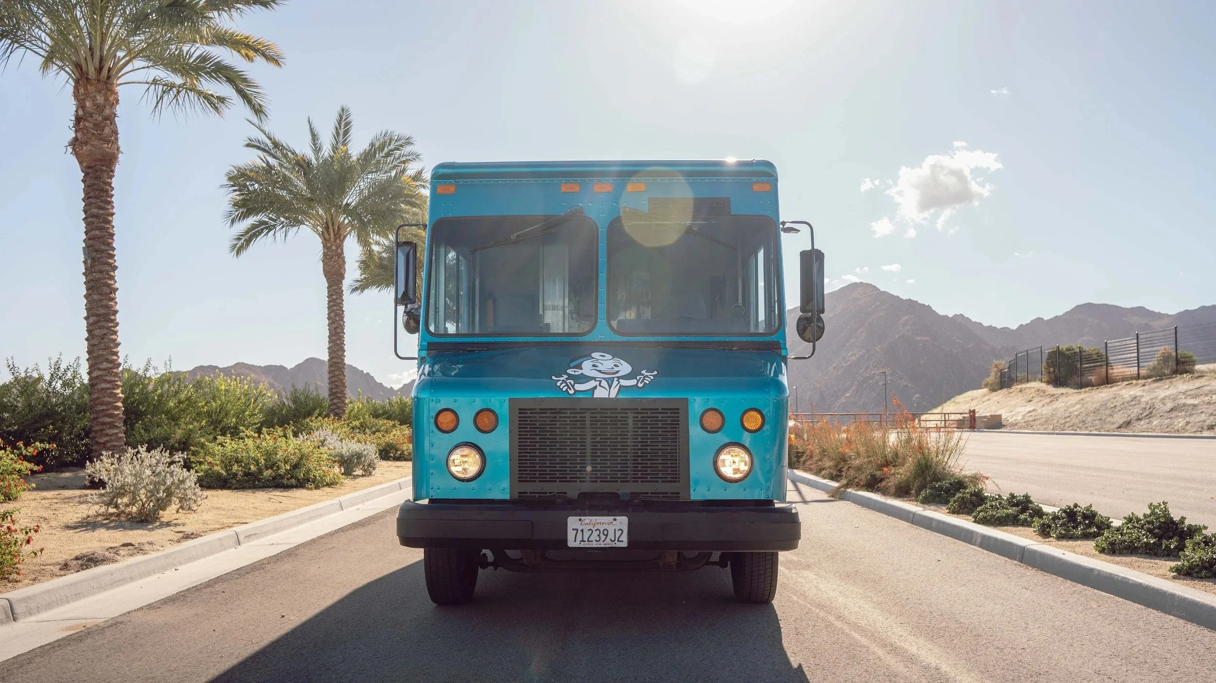 Blue mobile bike repair truck with logo of a smiling Dr. Bike on the front, parked in a sunny desert landscape with palm trees and mountains in the background.