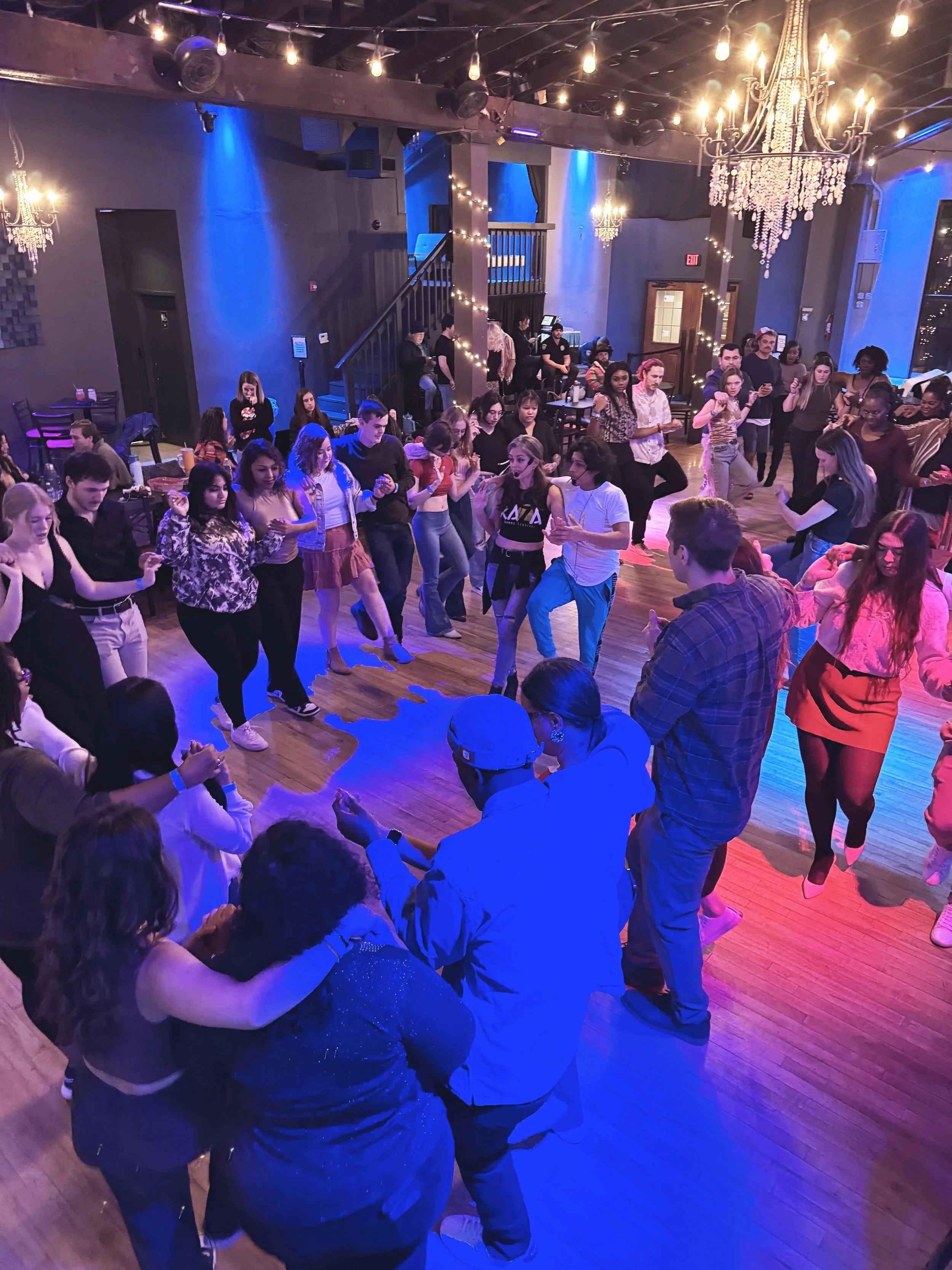 Group of people dancing in a dimly lit room at Twenty Seven Venue with blue and pink lighting, chandeliers, and a wooden floor, engaging in an organized circle dance.