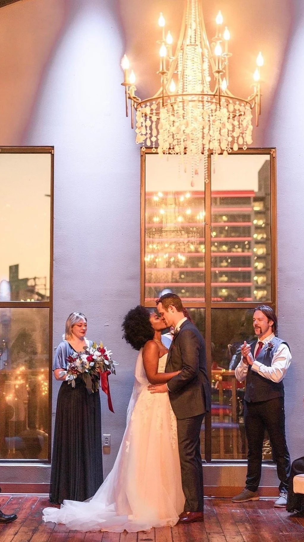 Bride and groom kissing at wedding ceremony with bridesmaid holding bouquet and groomsman applauding, under a chandelier.