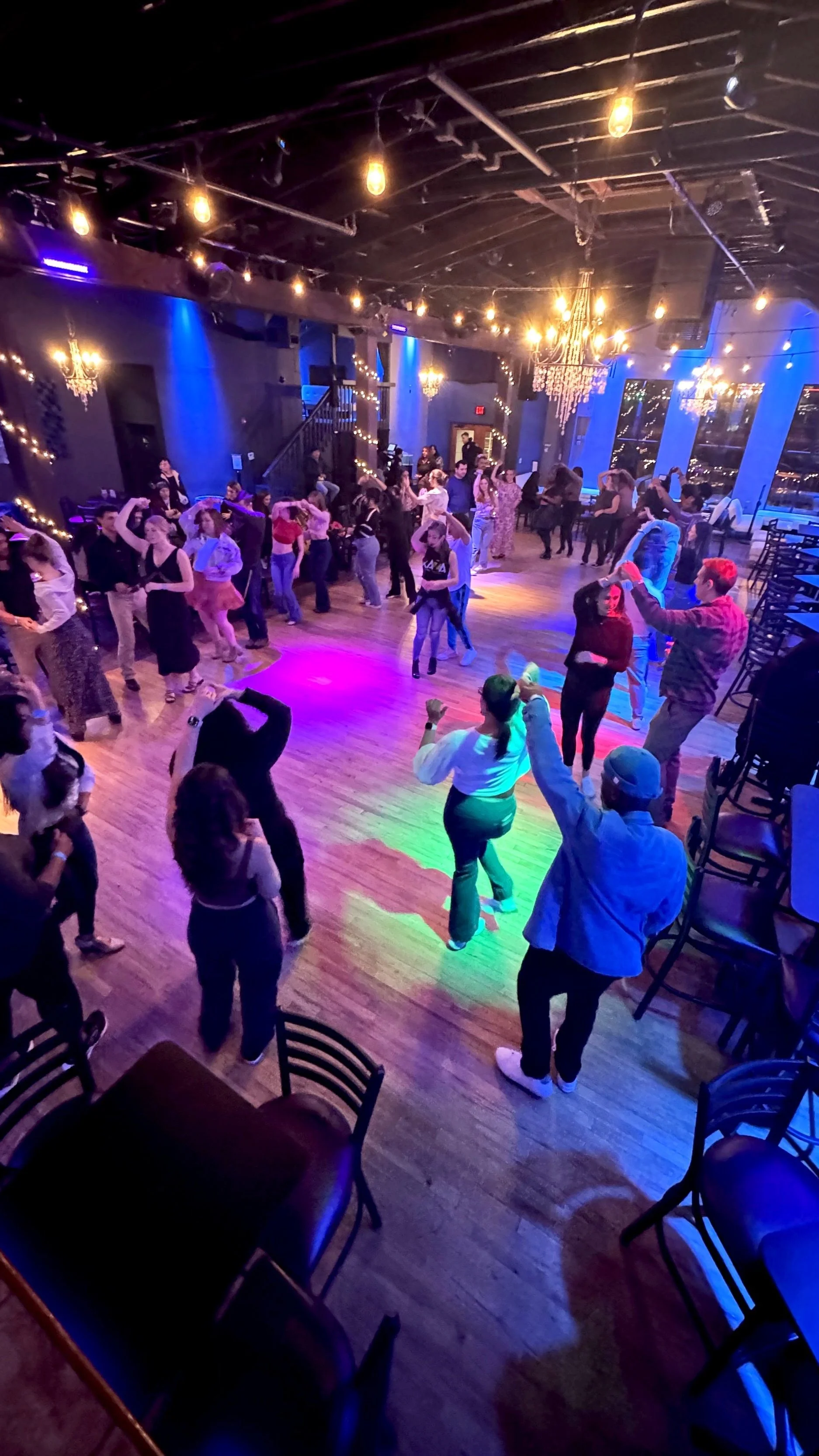 A lively indoor dance event with people enjoying partner dancing, under warm decorative lighting in a spacious venue.