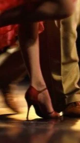 Close-up of two people dancing, showing legs and shoes, with a woman wearing red high heels and a man wearing beige pants and brown shoes.