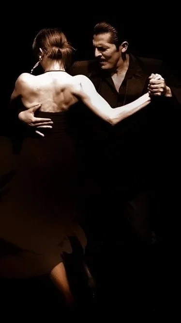 A couple performing a tango dance in a dimly lit setting. The man is dressed in black and the woman wears a dark dress, holding each other in a passionate embrace.
