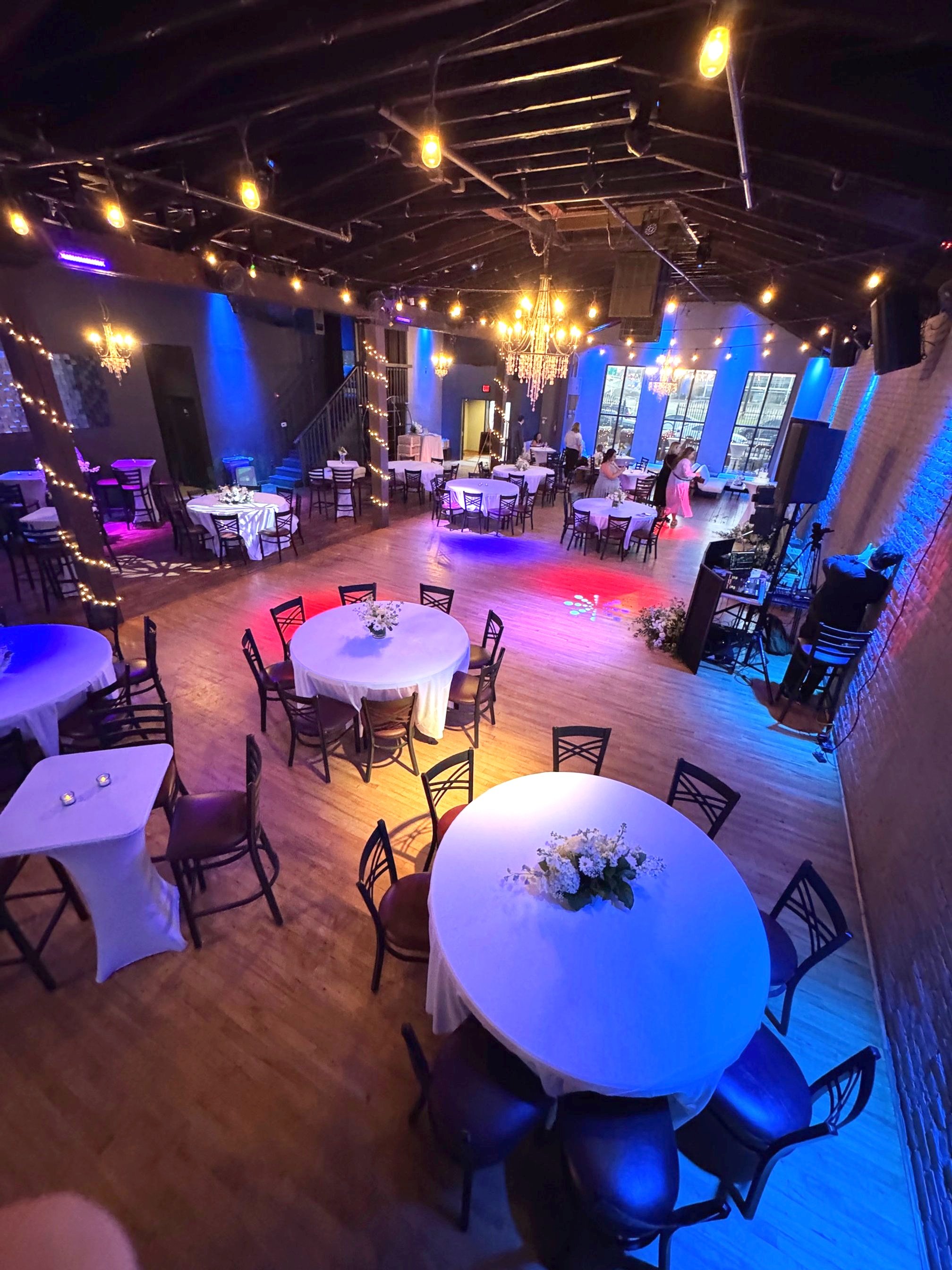 Elegant event venue in downtown Little Rock with round tables draped in white linen for a wedding, decorated with floral centerpieces. Dim lighting with chandeliers and string lights create a cozy atmosphere.