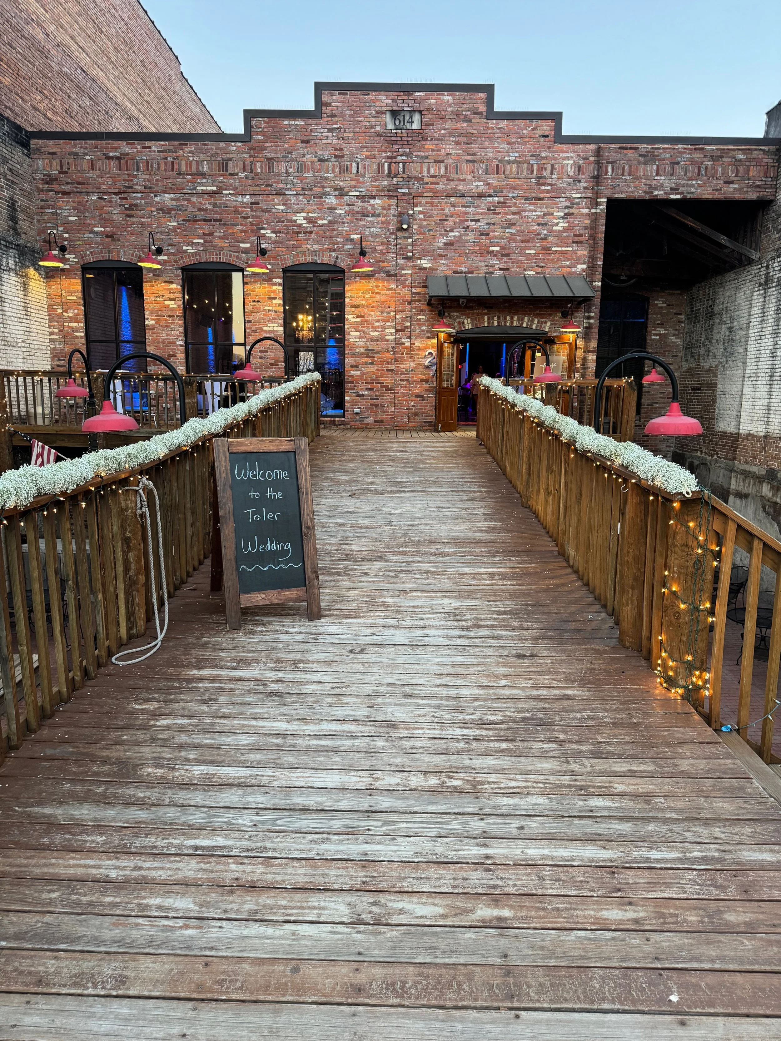 Twenty Seven event venue's front showing it is a brick building with wooden walkway leading to entrance, decorated with string lights and flowers for a wedding reception in downtown Little Rock