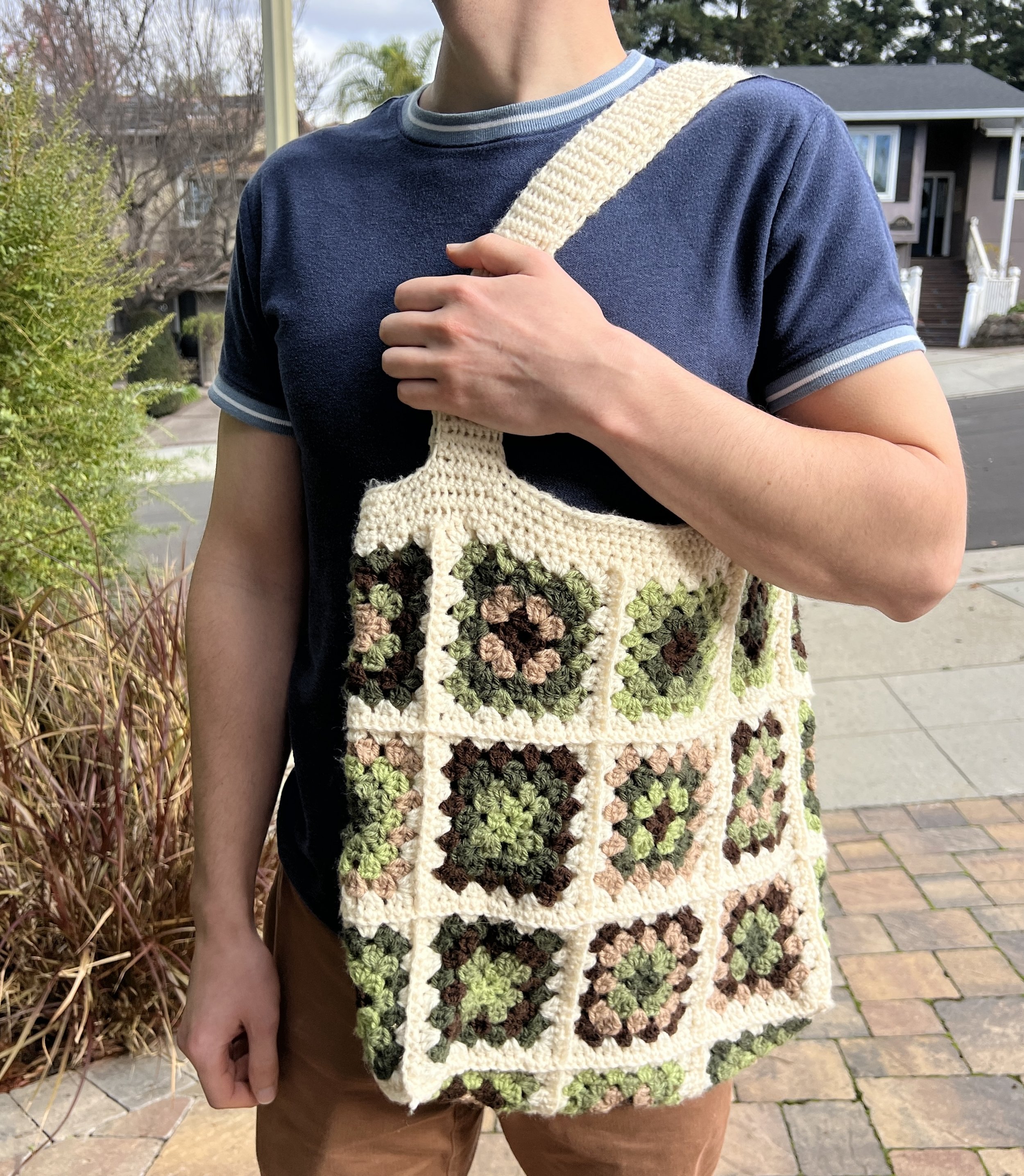 A person holding a crocheted bag with a granny square pattern featuring green, brown, and pink colors.