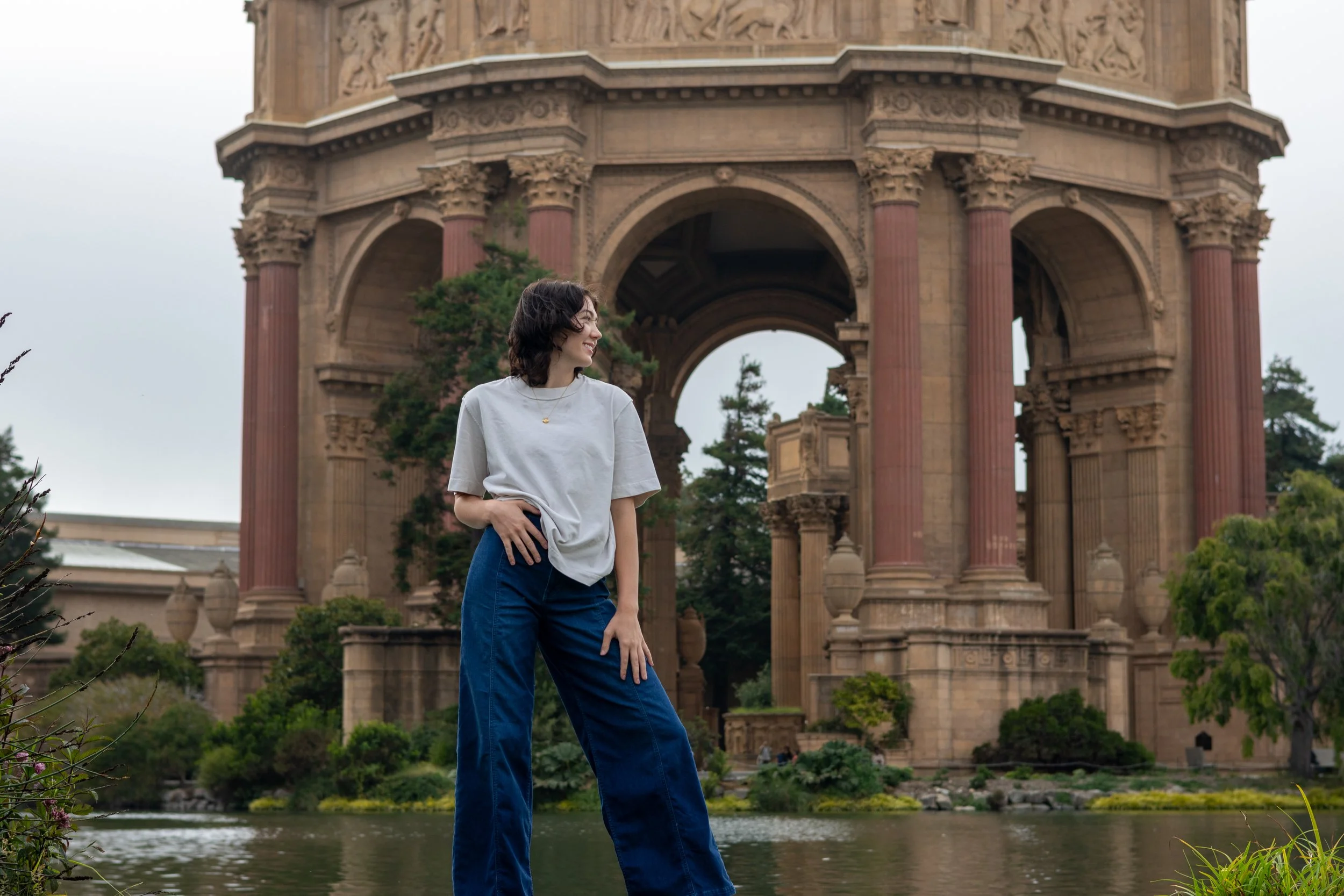 A woman with short dark hair standing near a body of water, smiling and looking to her left, with a historic stone archway structure and trees in the background.