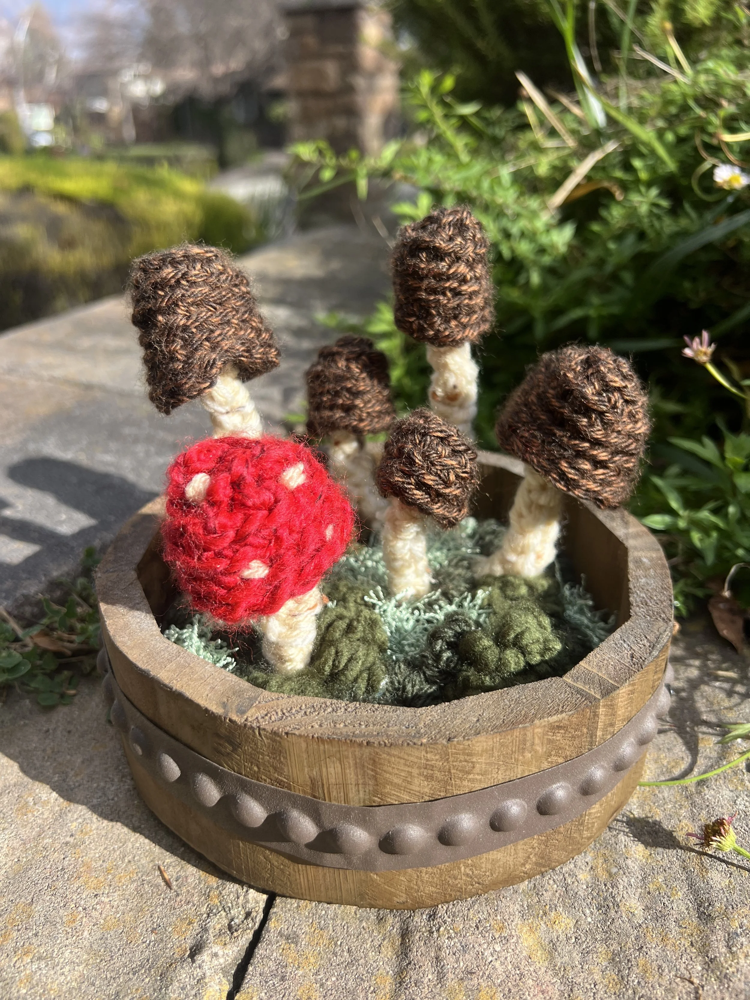 Felted mushroom garden in a wooden bowl with green moss and foliage, outdoors on a sunny day.