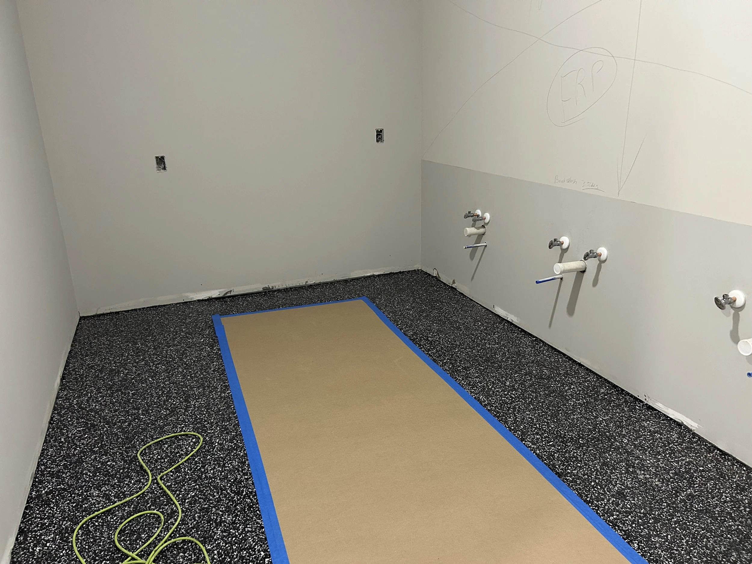 Room in the Crucible Fit buildout with unfinished walls, exposed plumbing, and floor markings.