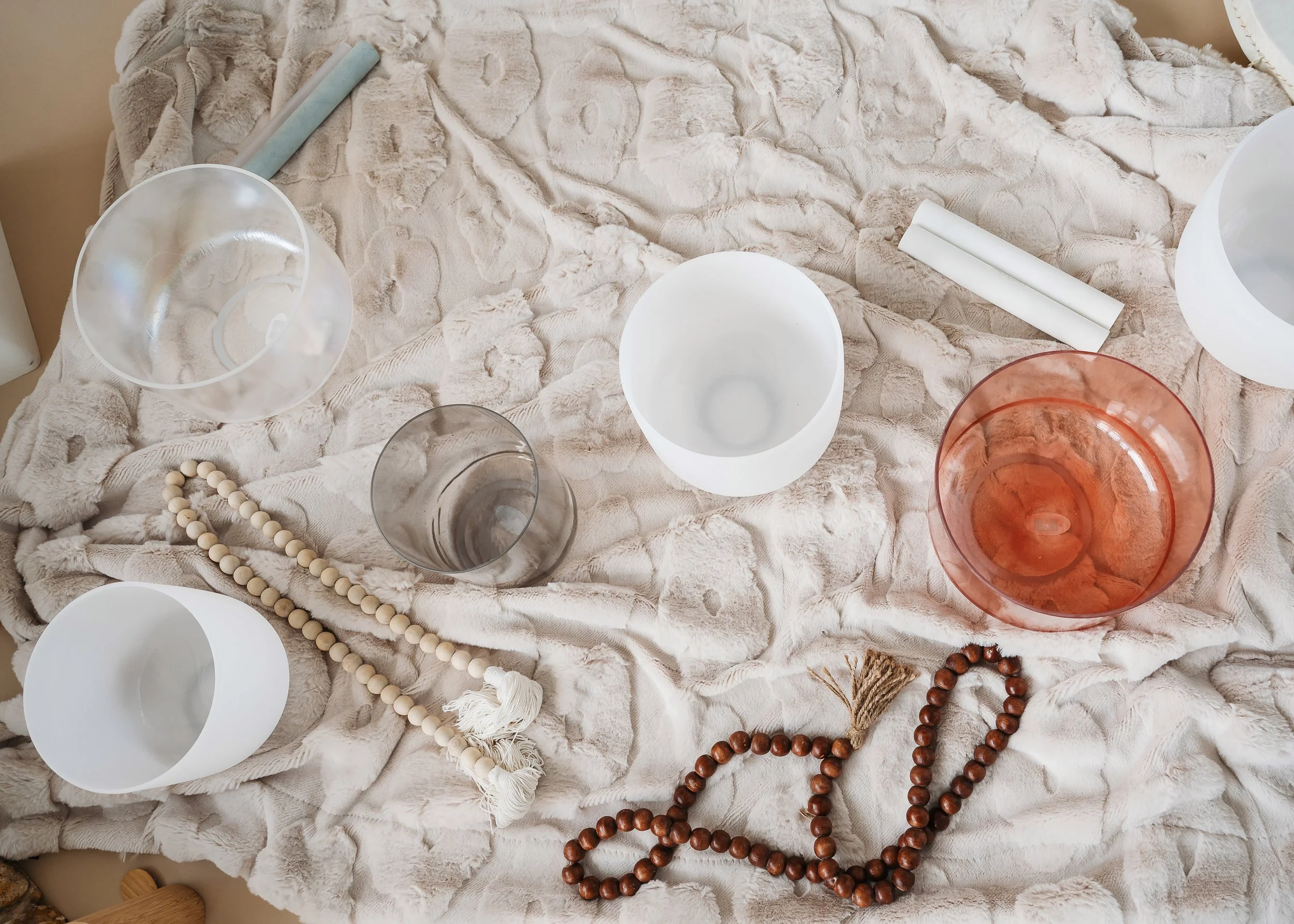 A soft, beige, plush blanket with decorative wooden beads scattered on top, clear and frosted glass bowls, and rolled white paper placed on it.