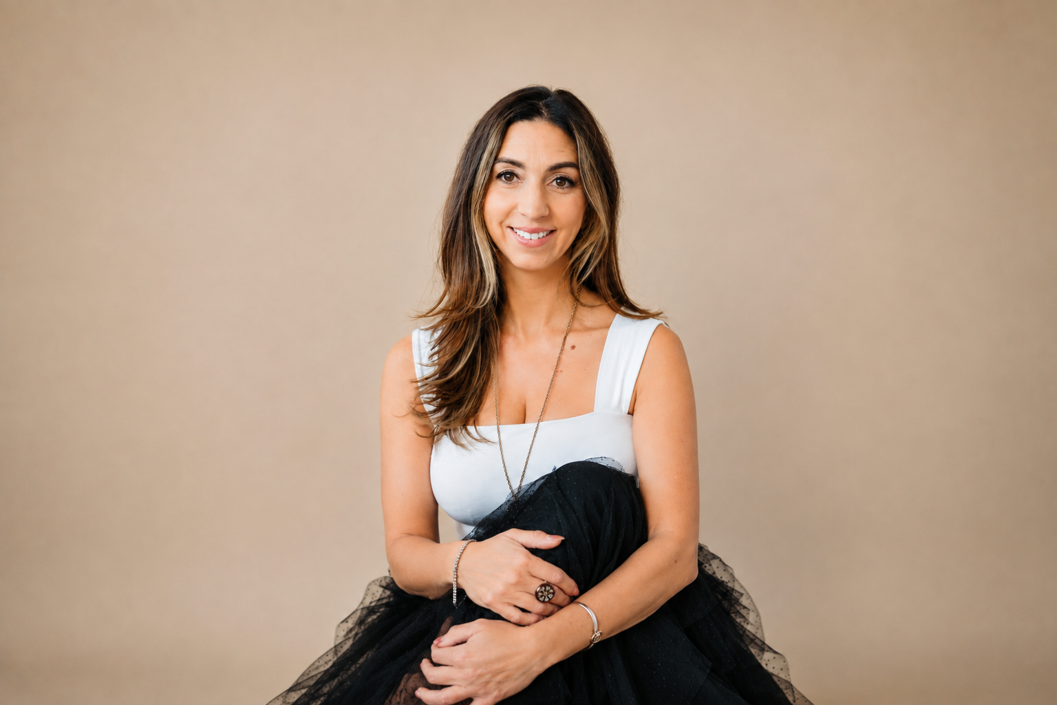 A smiling woman with long brown hair, wearing a white sleeveless top, black skirt, and jewelry, sitting against a plain beige background.