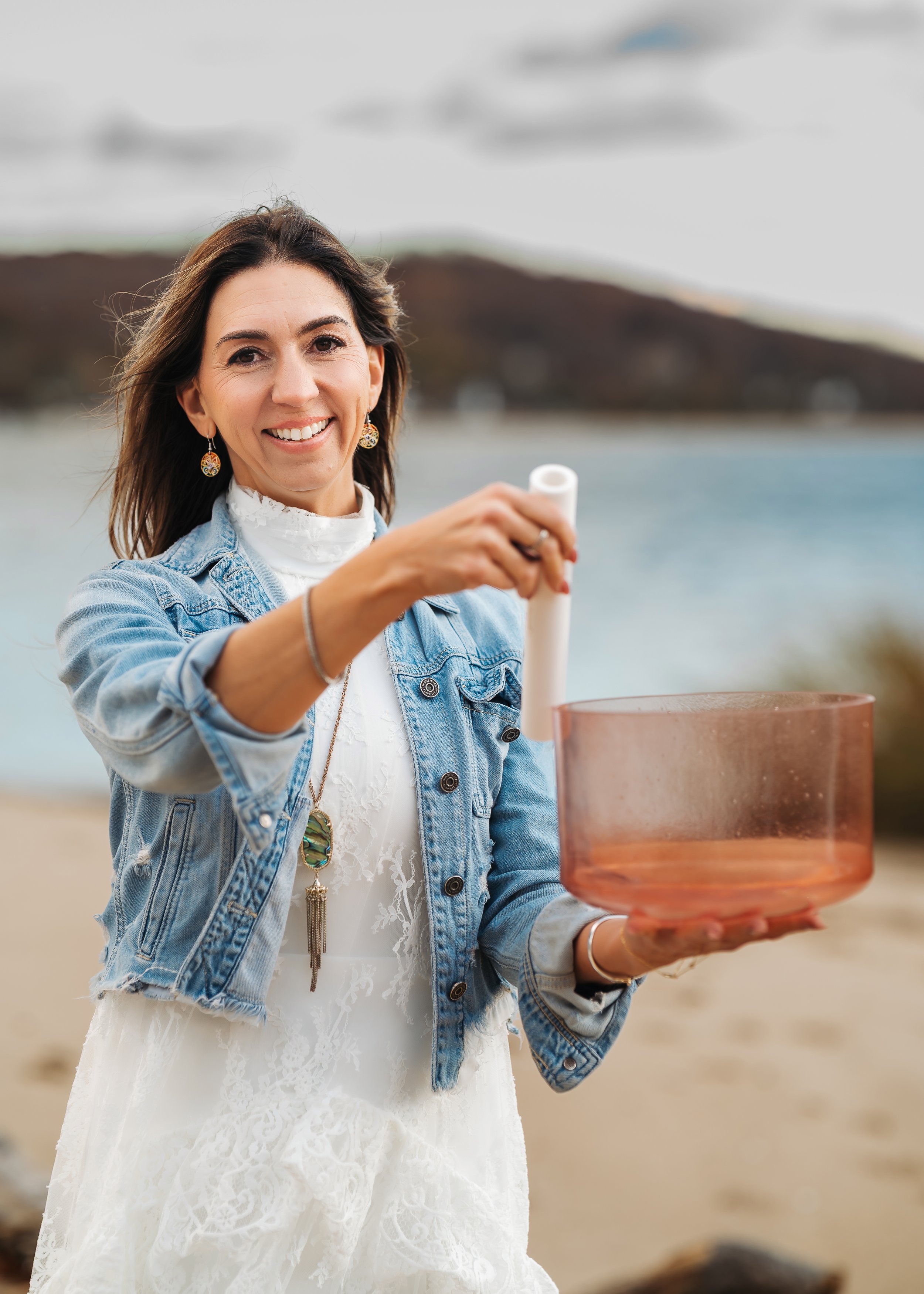 A smiling woman in a white dress and denim jacket holding a large pink glass bowl and a rolling pin outdoors near water with cloudy skies.