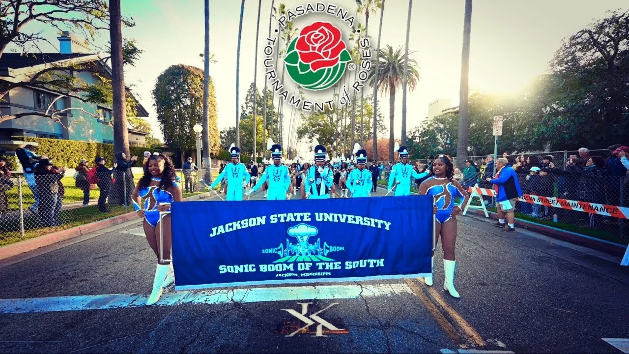A parade with drumline performers from Jackson State University in blue uniforms and cheerleaders holding a banner for Jackson State University's Sonic Boom of the South. Spectators line the street behind a fence, with palm trees and buildings in the background during a sunny day.