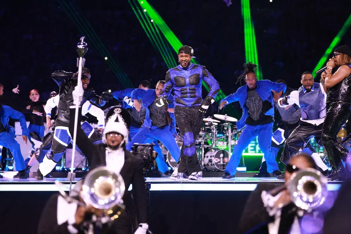 A lively stage performance with dancers in blue and black costumes, a man in the center wearing a glittery blue and black outfit, and a singer on the right holding a microphone, with green laser lights in the background.