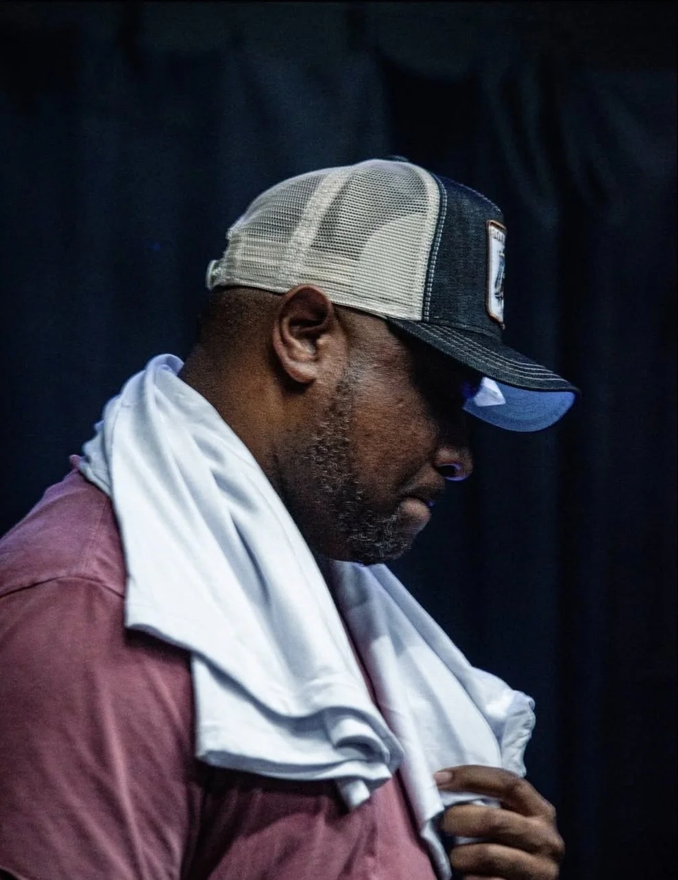 Side profile of a man wearing a trucker cap and a towel draped over his shoulders, with a dark background.