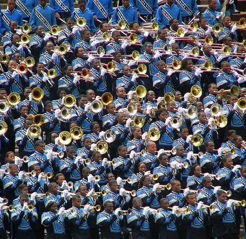 A large group of marching band members dressed in blue and black uniforms playing brass instruments like trumpets and trombones during a parade.