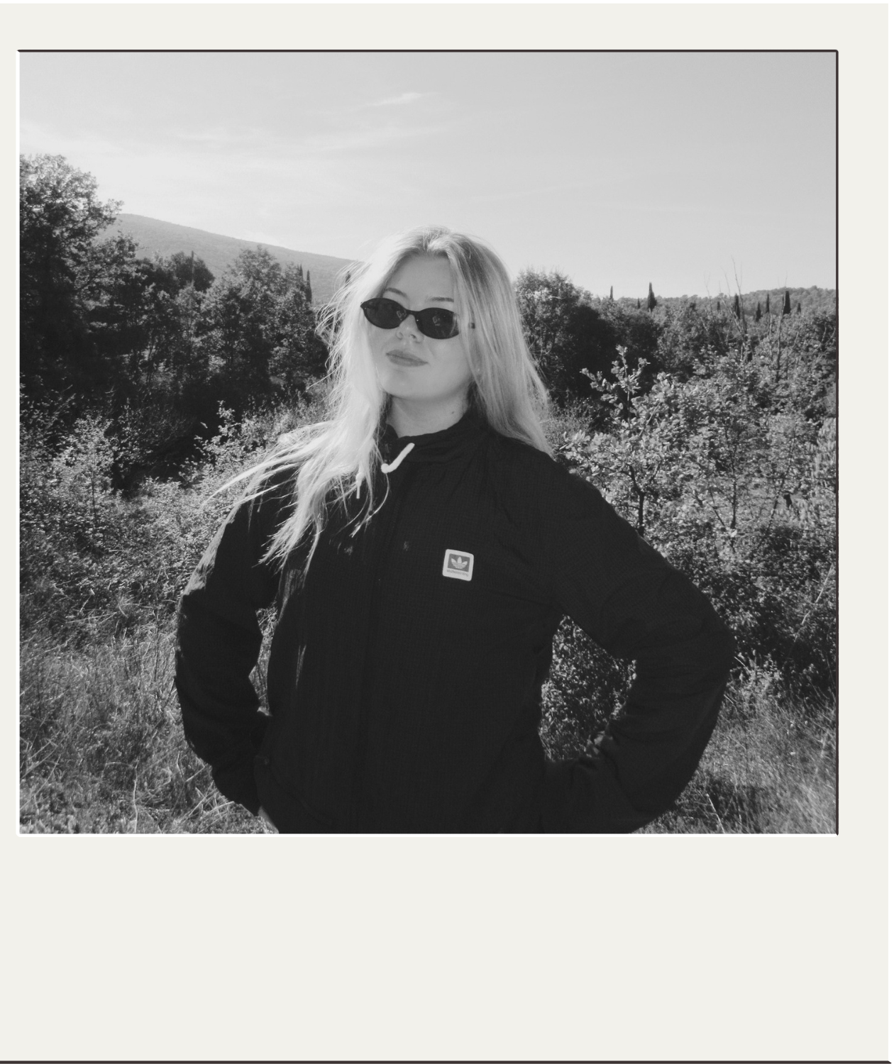 Black-and-white portrait of a woman wearing sunglasses and an Adidas jacket outdoors in a wooded landscape.