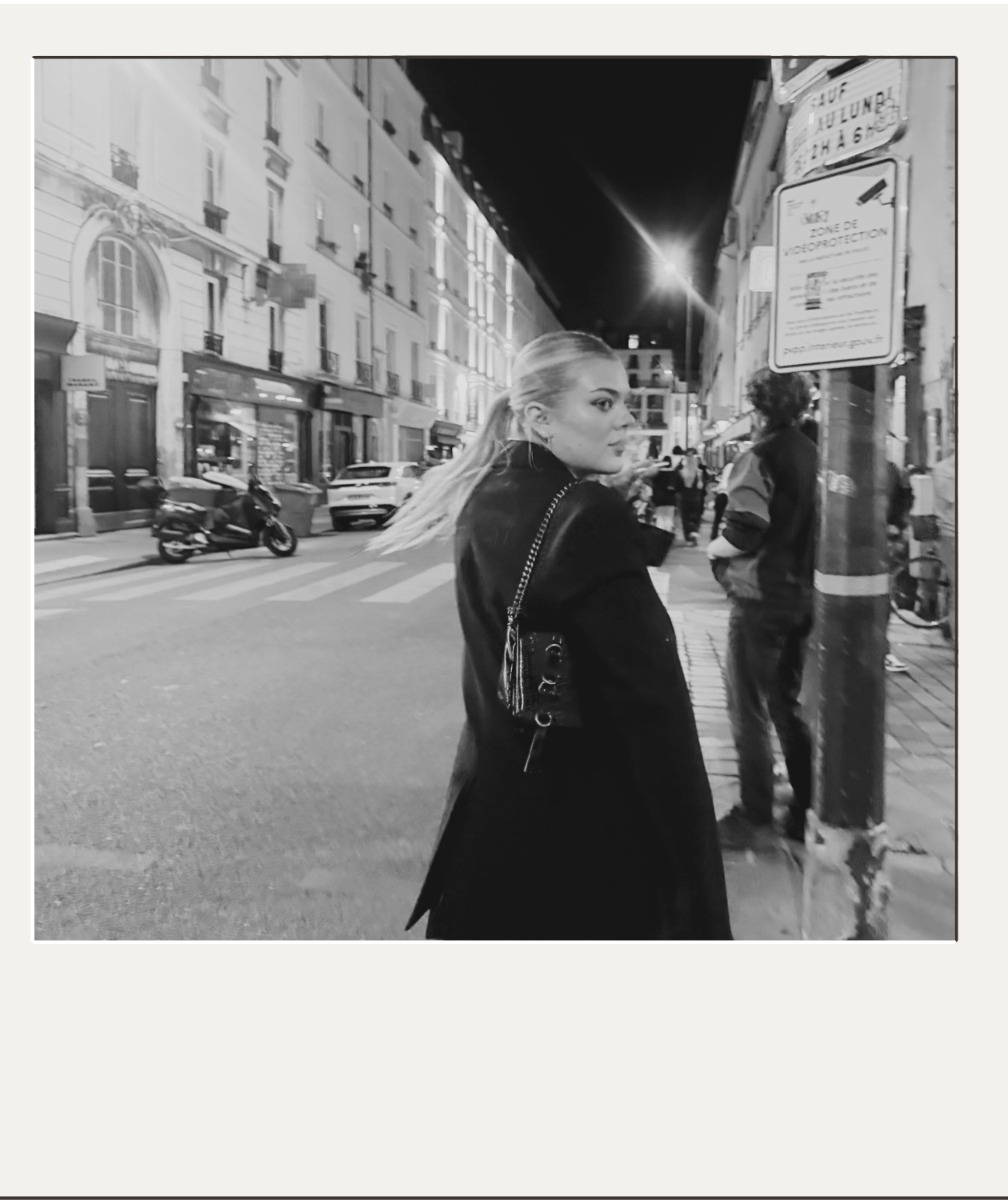 Black-and-white photo of a woman with a ponytail and shoulder bag walking along a city street at night.