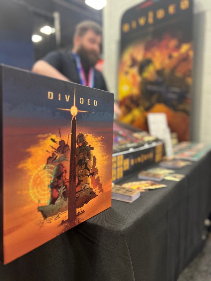 A tabletop display of a board game titled "Divided" featuring artwork of soldiers and a sunset, with a person in the background at Origins convention.