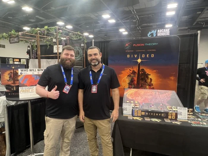 Joey And Kenneth co-designer of Divided stand smiling in front of a booth for the game 'Divided' at a convention, with game artwork, promotional displays, and other attendees in the background.