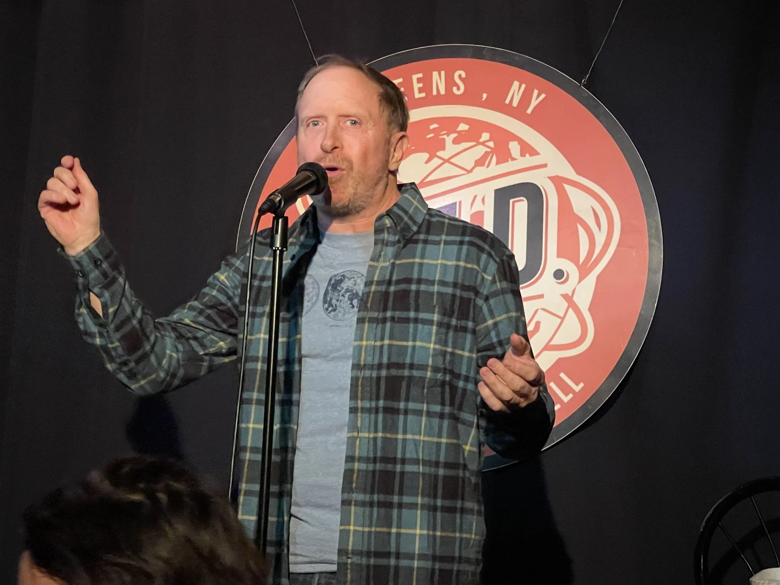 William in a plaid shirt and graphic t-shirt speaking into a microphone on stage at a storytelling performance venue, with a round logo in the background that reads 'QED".