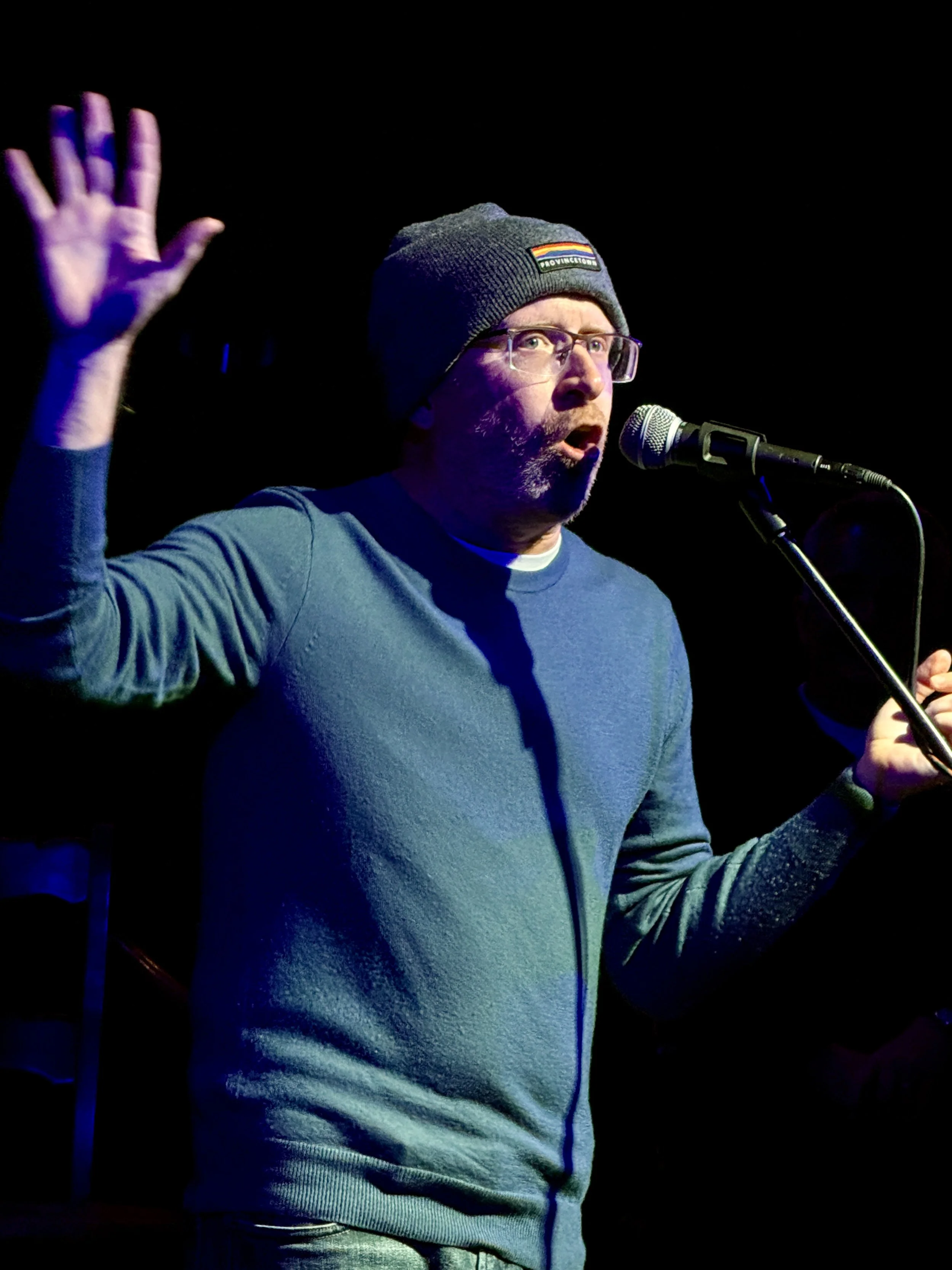 William wearing a beanie cap, glasses, and a blue sweater speaking into a microphone with one hand raised in the air.