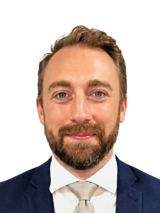Headshot of a man with light brown hair, a beard, and blue eyes, wearing a dark suit, white shirt, and beige tie, smiling against a black background.