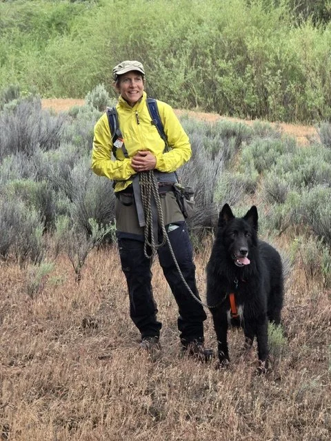 A woman hiking outdoors with a black German Shepherd dog. She is wearing a yellow jacket, hat, and carrying a backpack. The landscape has bushes and grassy terrain.