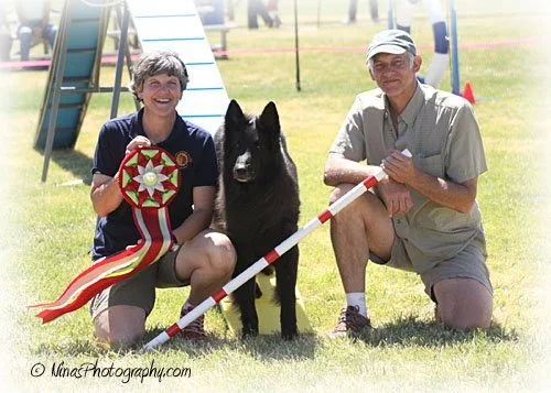 A woman and a man kneeling on grass with a black German Shepherd dog, holding a colorful ribbon and a barrier pole, at an outdoor event.