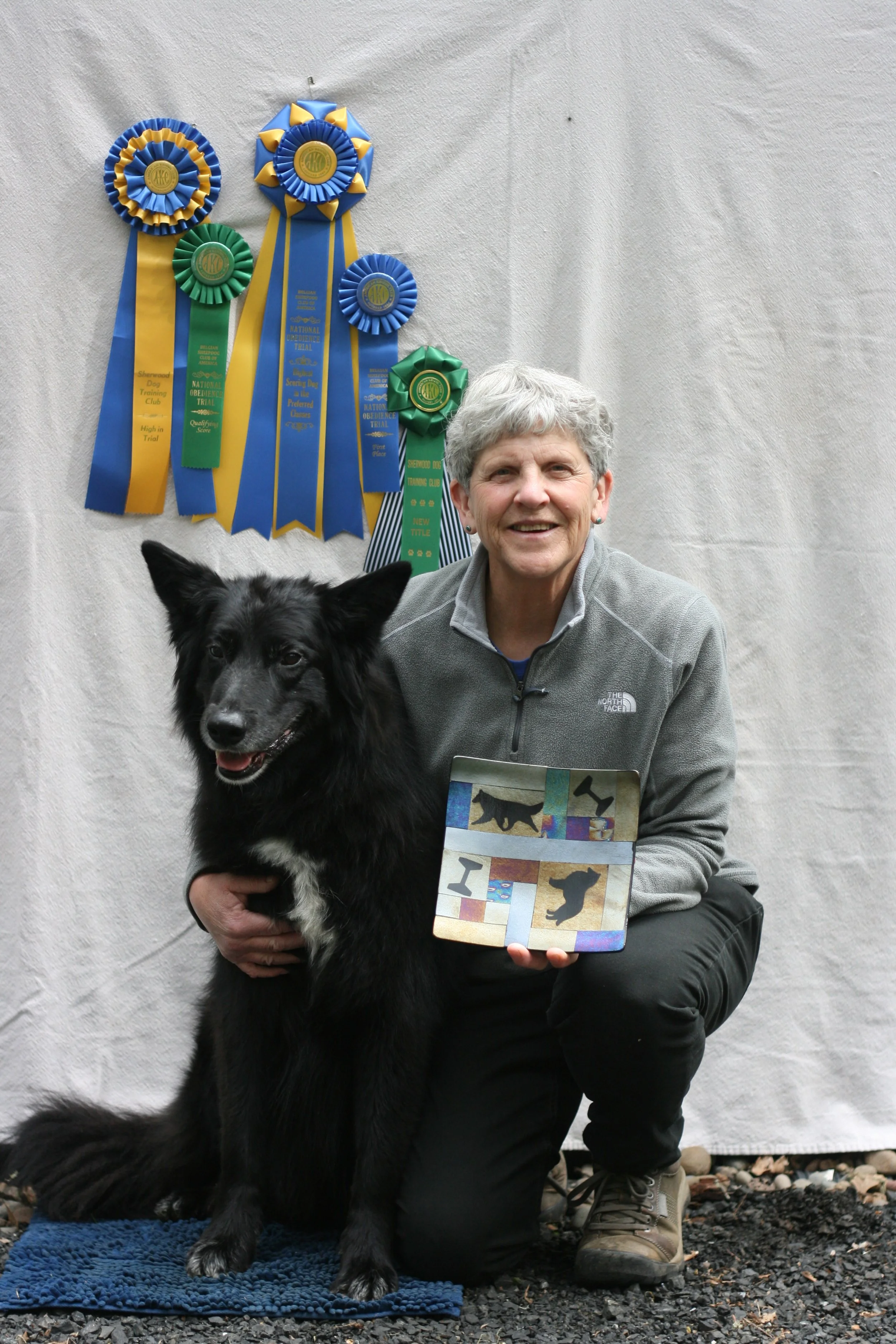A woman with short gray hair and a young black and white dog are posing for a photo. The woman is kneeling on one knee, holding a colorful abstract art piece. The dog is standing on a blue textured mat, looking forward with its mouth slightly open. B