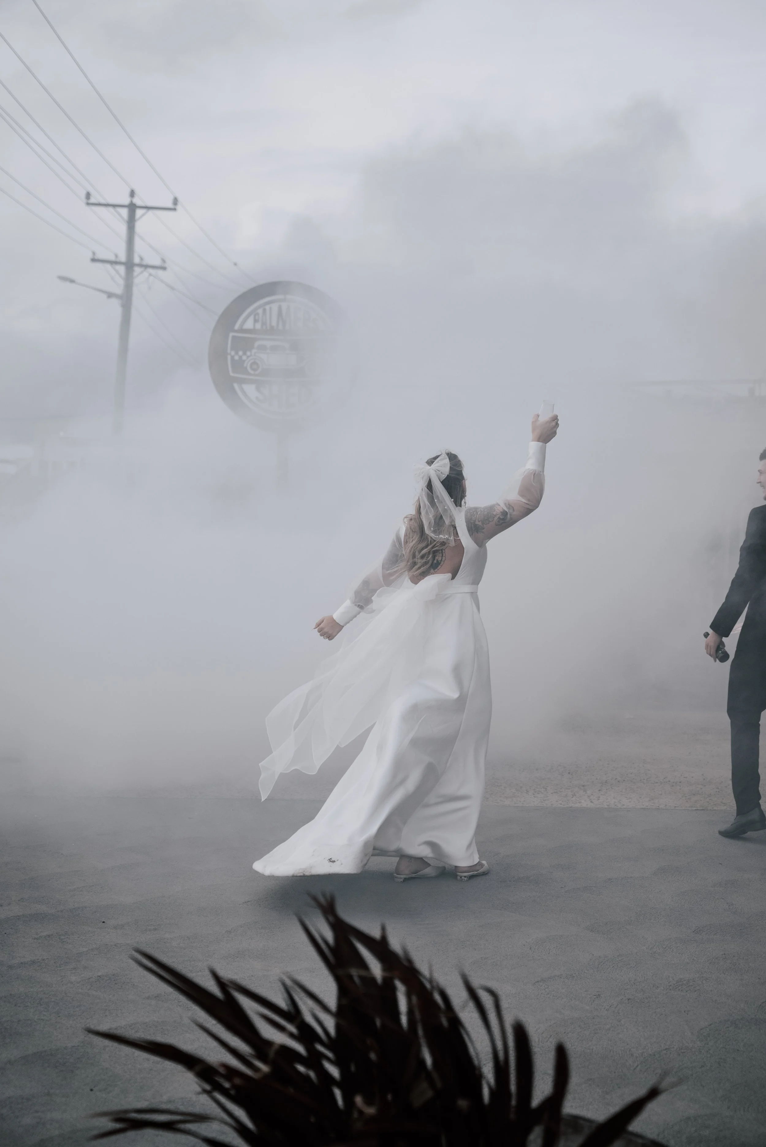 A bride in white with a hair bow raises her wine glass in a cloud of burnt rubber from a car doing a burnout