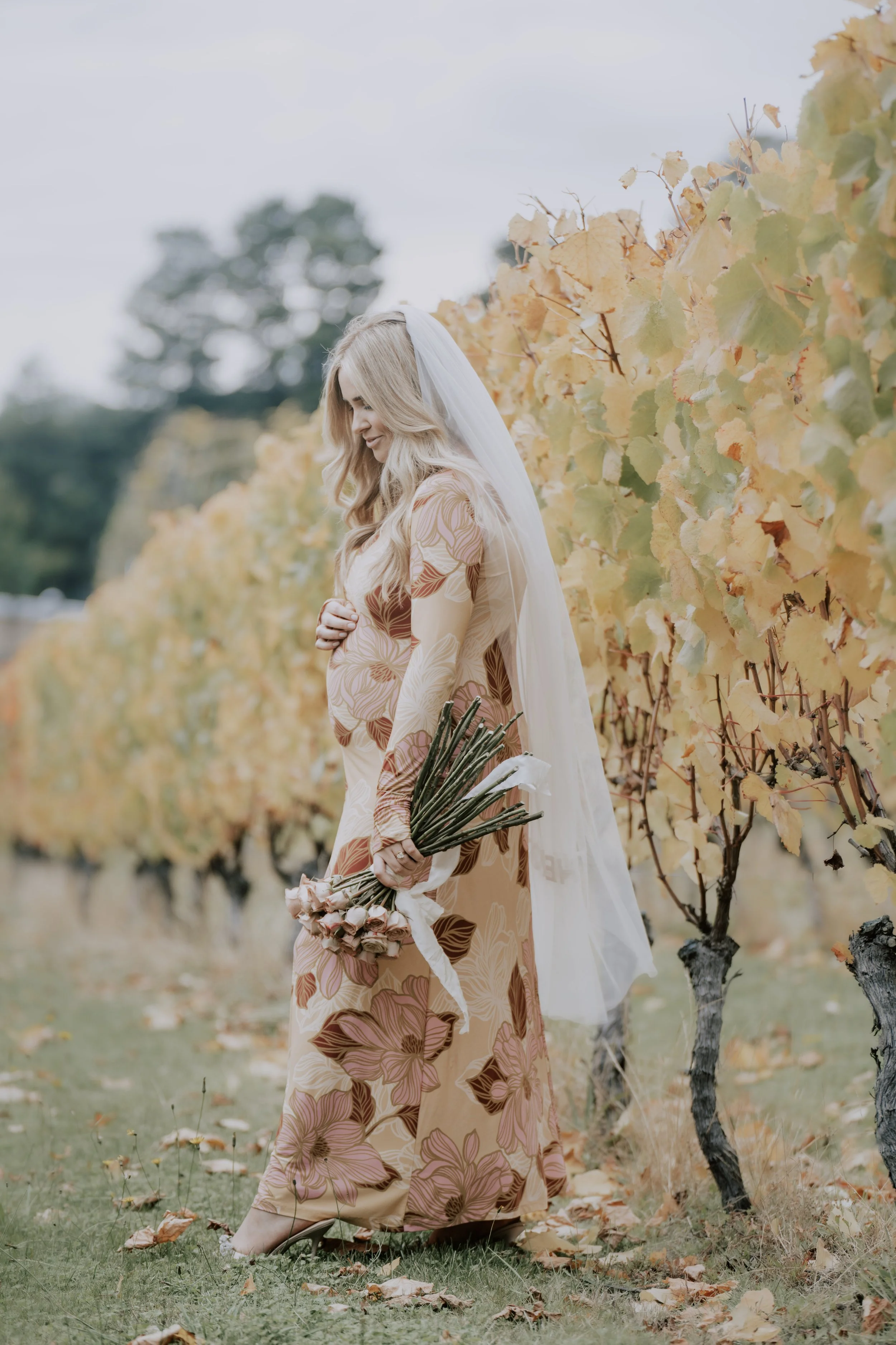 A bride with a long veil smiles down at her pregnant belly while she stands in a vineyard in a pink and orange wedding dress