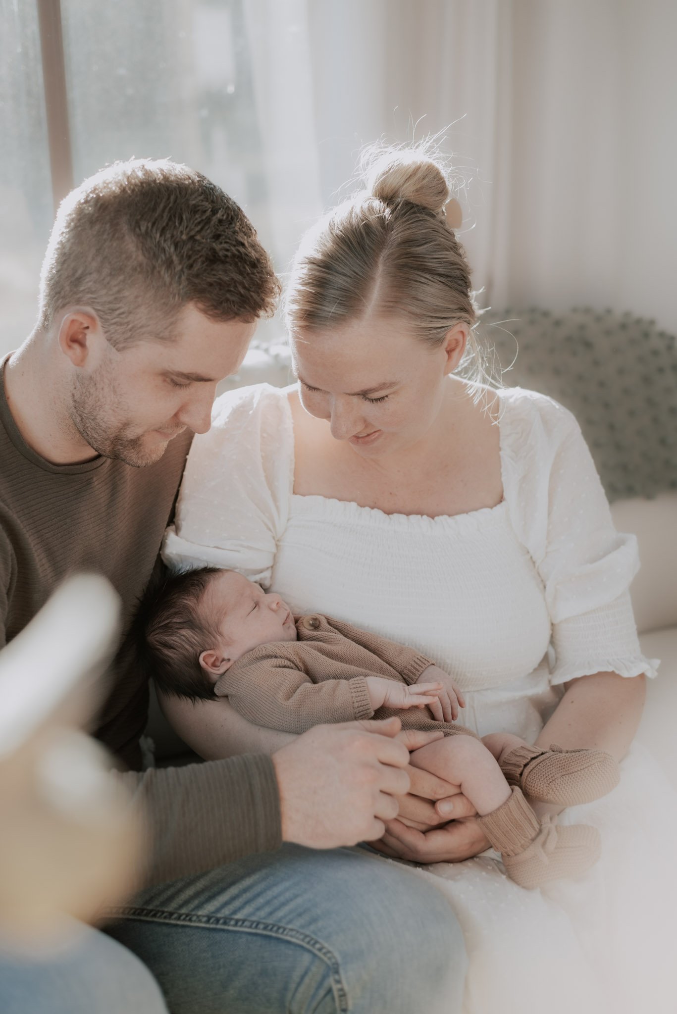 A young couple with light skin looking lovingly at their sleeping baby, who is cradled on their laps, indoors near a window.