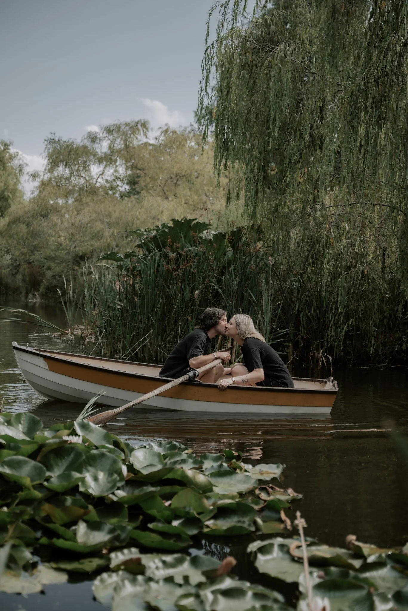 Two people share a kiss in a small wooden rowboat on a calm lake surrounded by tall trees and lush greenery.