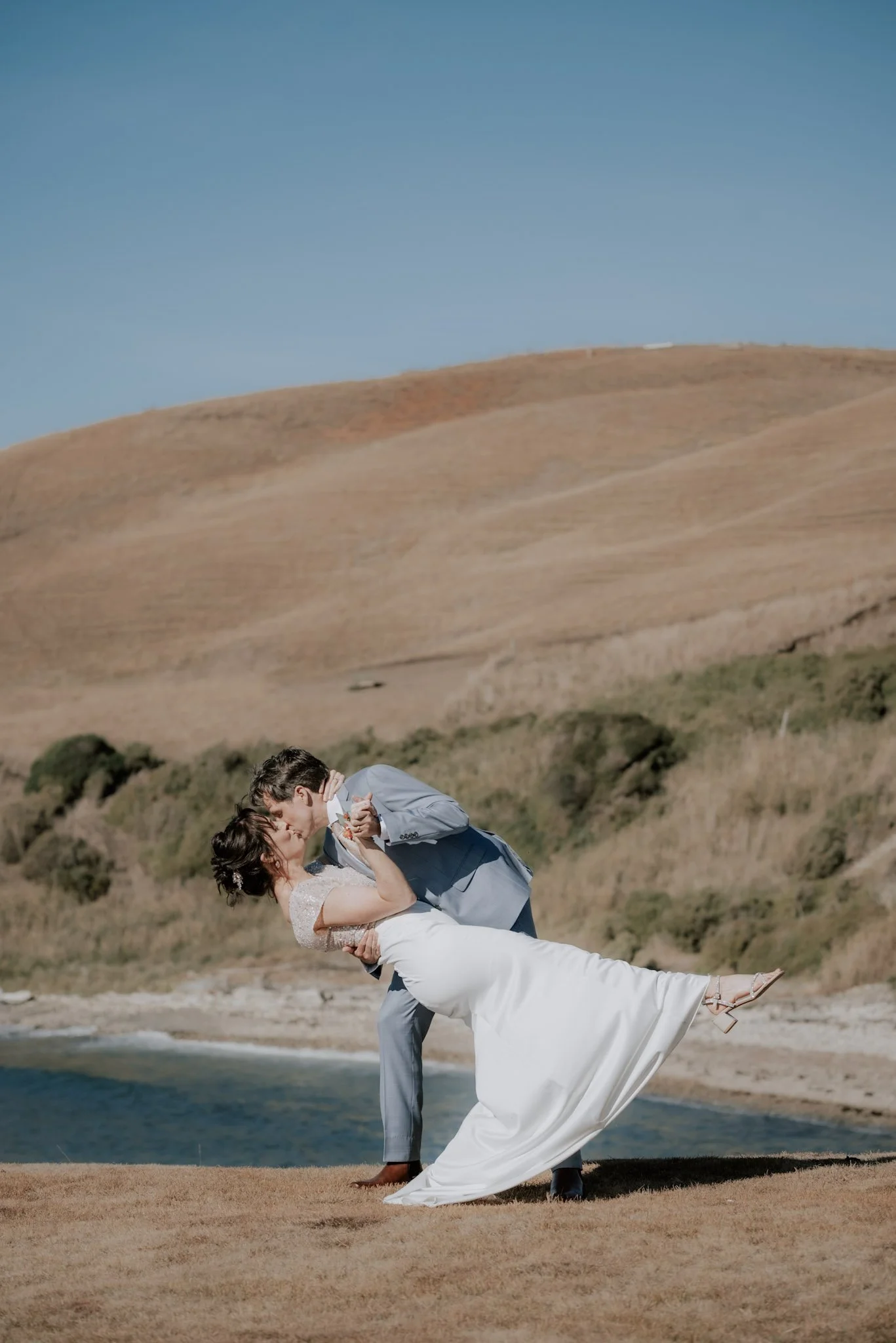 A bride and groom sharing a kiss outdoors near water, with hills in the background.