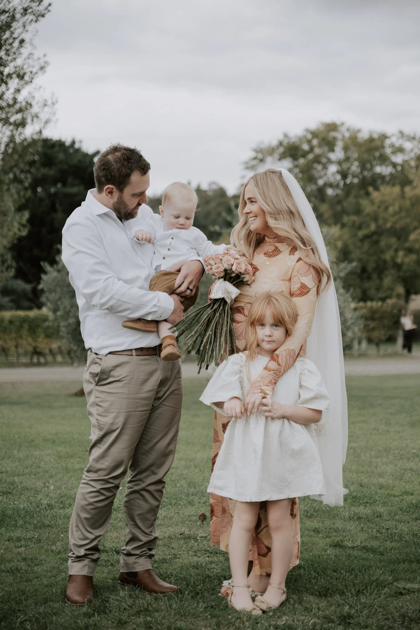 A family of four celebrating a wedding outdoors. The mother, wearing a floral dress and veil, holds a bouquet of pink roses. The father, in a white shirt and khaki pants, holds a young child. A little girl with red hair in a white dress stands in fro