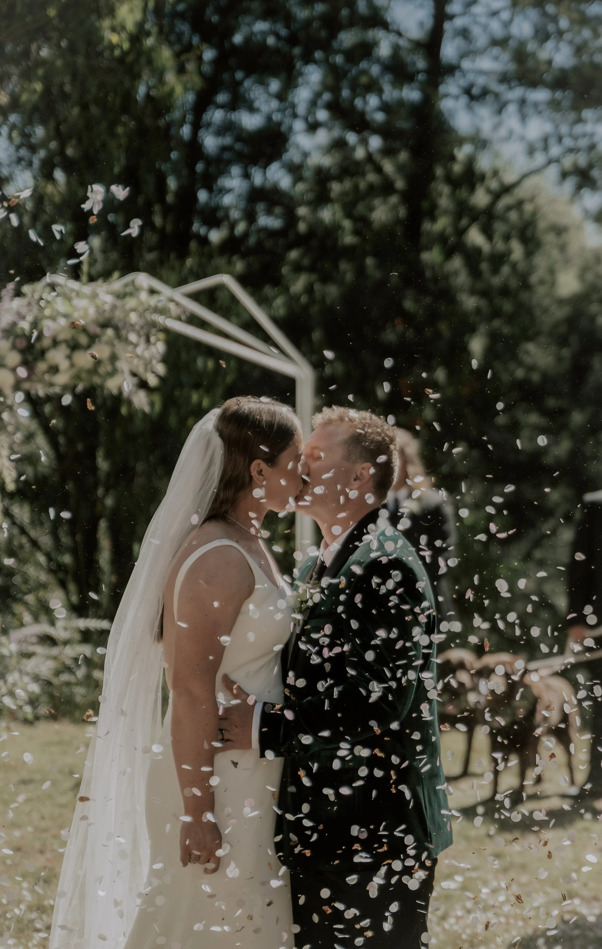 A bride and groom kissing at their outdoor wedding, surrounded by floating confetti and greenery.
