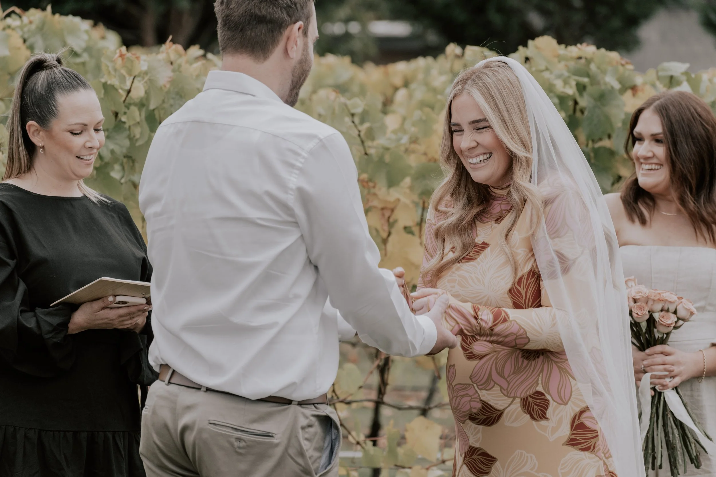 A wedding ceremony outdoors with a bride and groom holding hands, smiling, as they exchange vows, surrounded by a vineyard and bridesmaids.