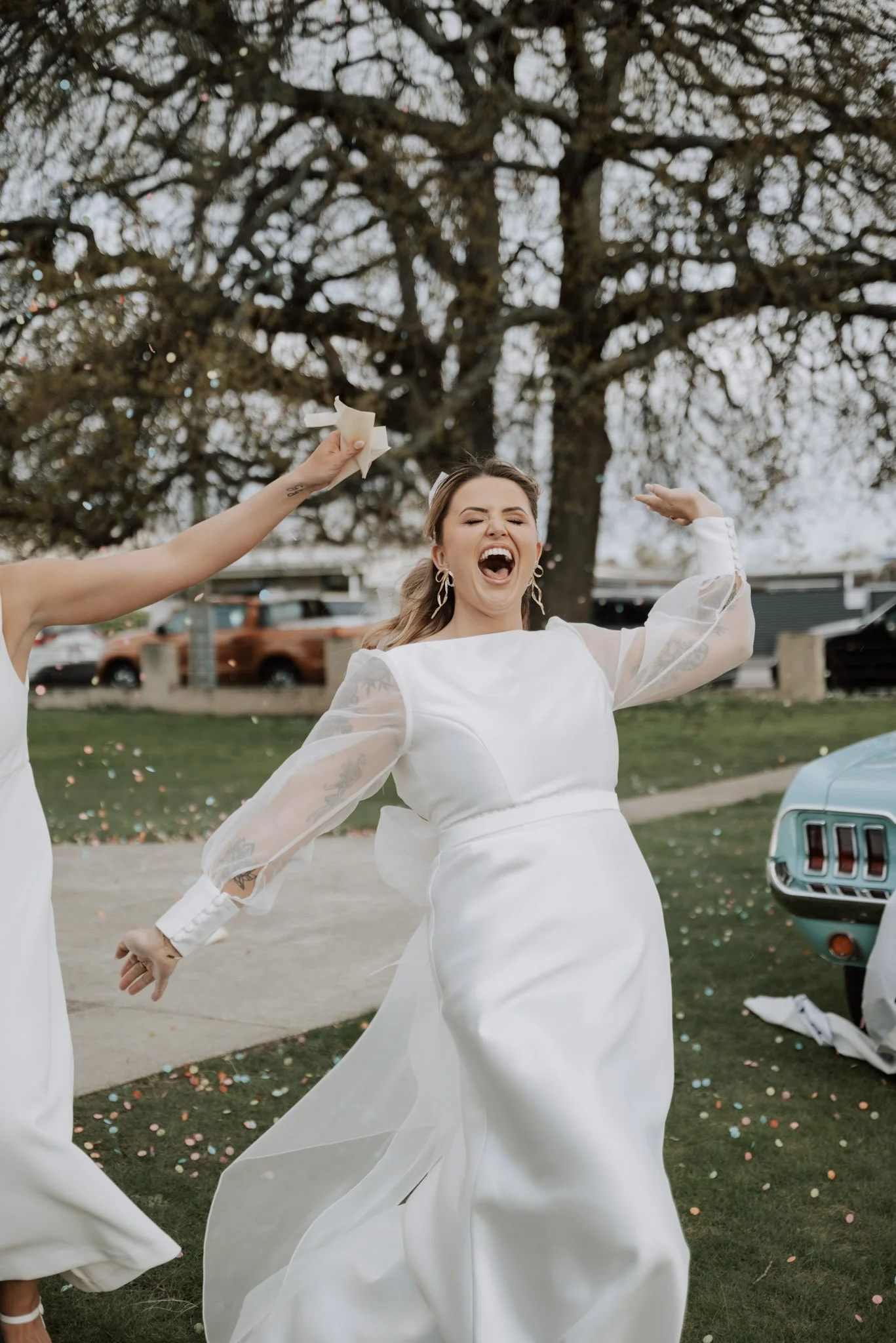 Bride in a white wedding dress celebrating outdoors with her wedding party, surrounded by confetti, with a large tree and a vintage car in the background.