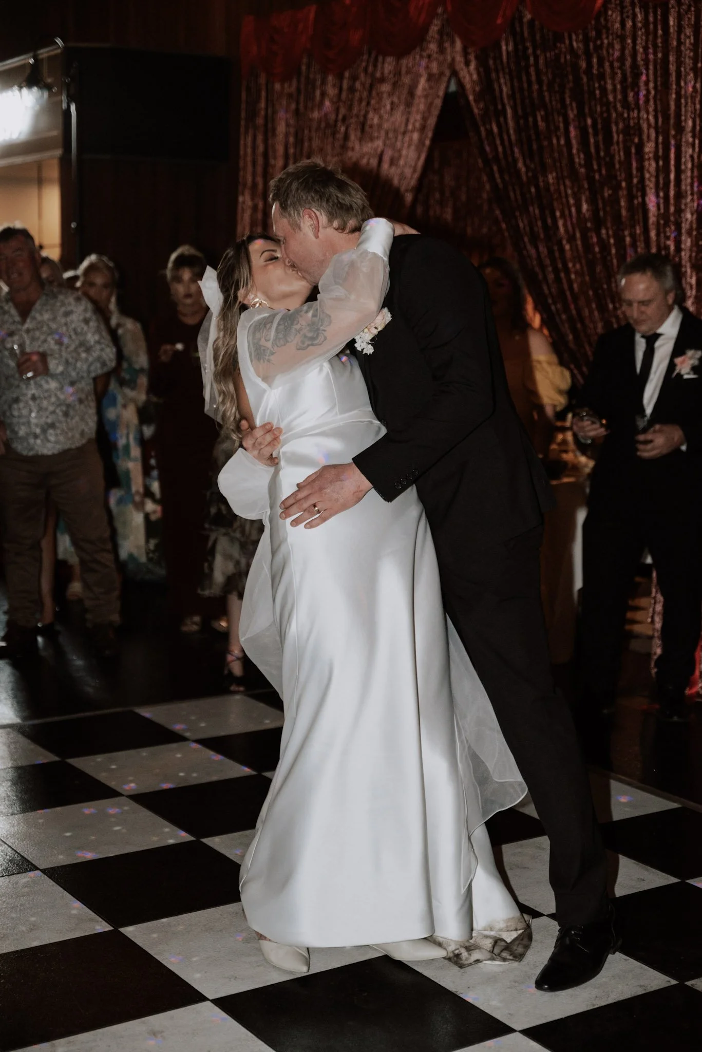 A bride and groom share a kiss during their wedding dance, with guests watching in the background.