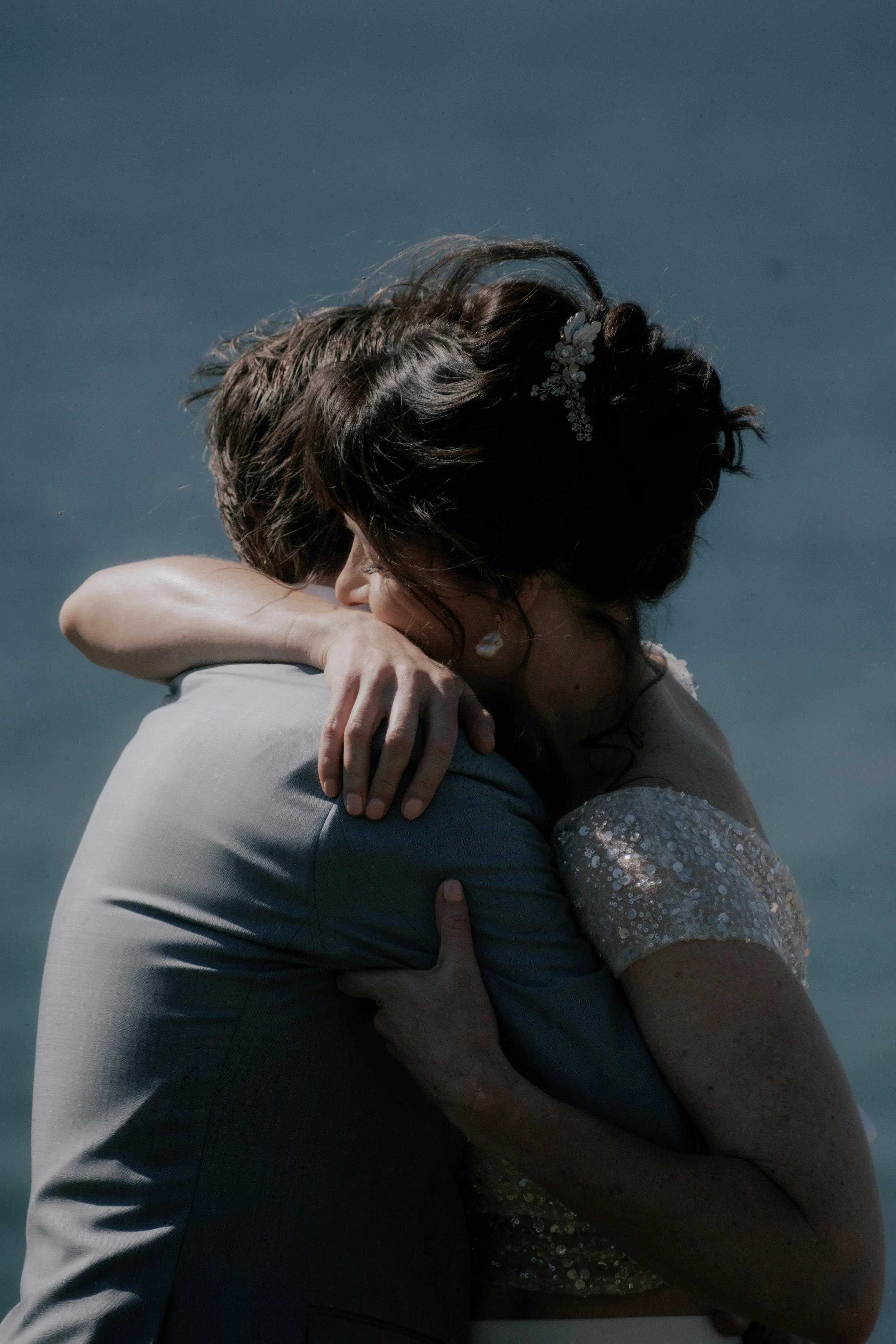 A bride and groom share a heartfelt embrace in wedding attire near a body of water.