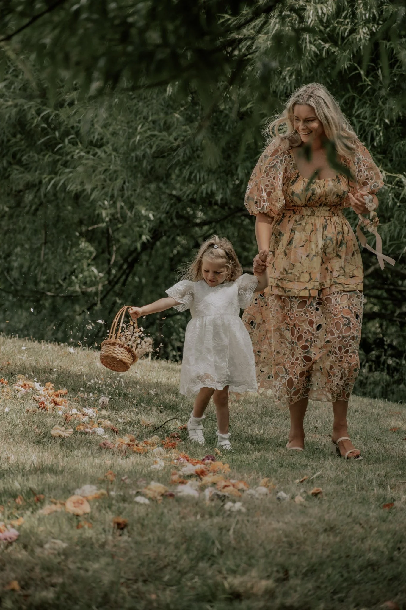 A woman and a small girl walk through a grassy area, surrounded by trees. The girl wears a white dress and carries a basket, scattering flowers on the ground. The woman wears a colorful, patterned dress and holds the girl's hand, both smiling.