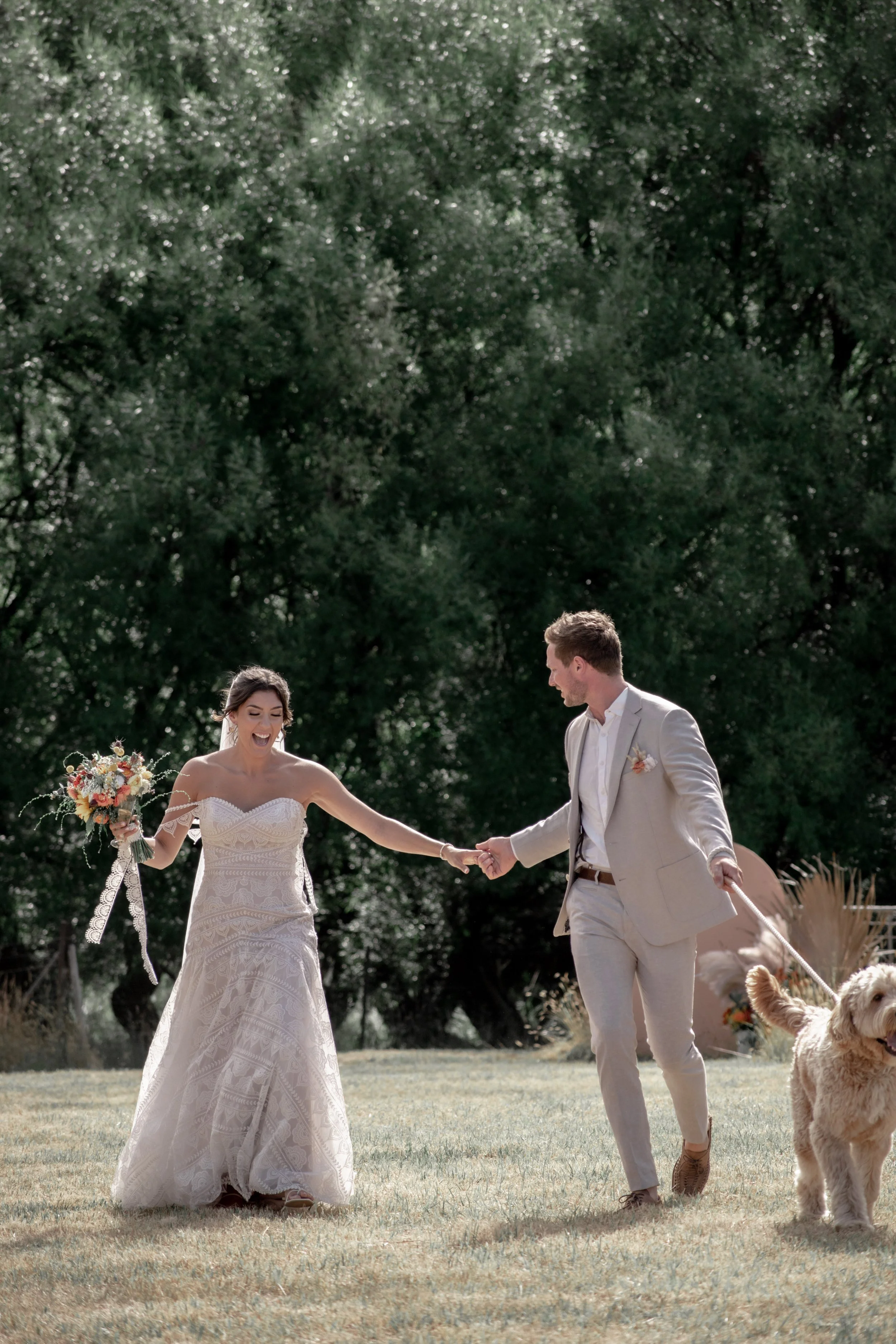 A bride and groom holding hands outdoors during a wedding ceremony, with a dog on a leash nearby. The bride is wearing a white lace wedding dress and holding a bouquet of flowers, while the groom is in a light-colored suit. They appear joyful and are