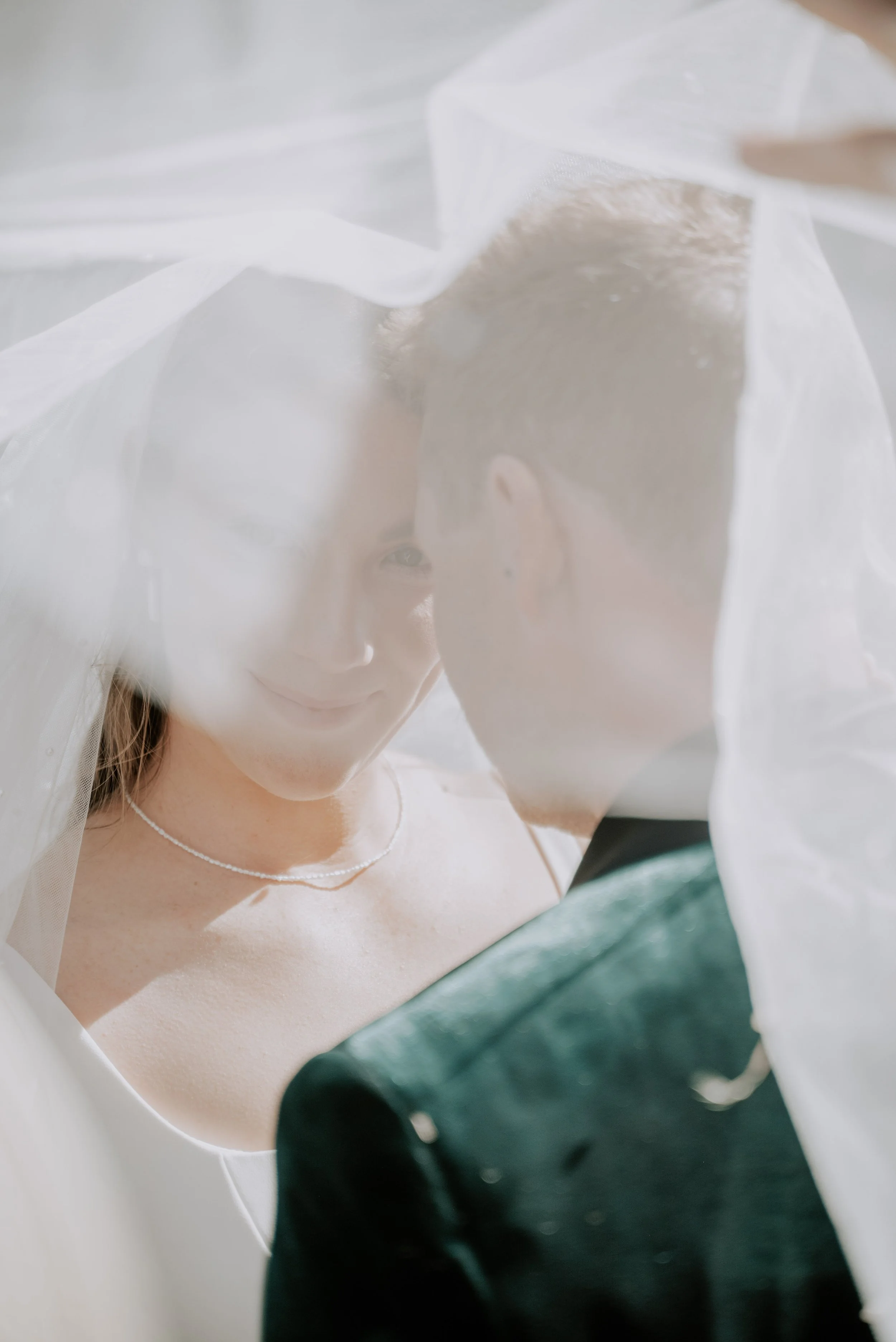 Bride and groom face each other under a veil, with the bride smiling softly.