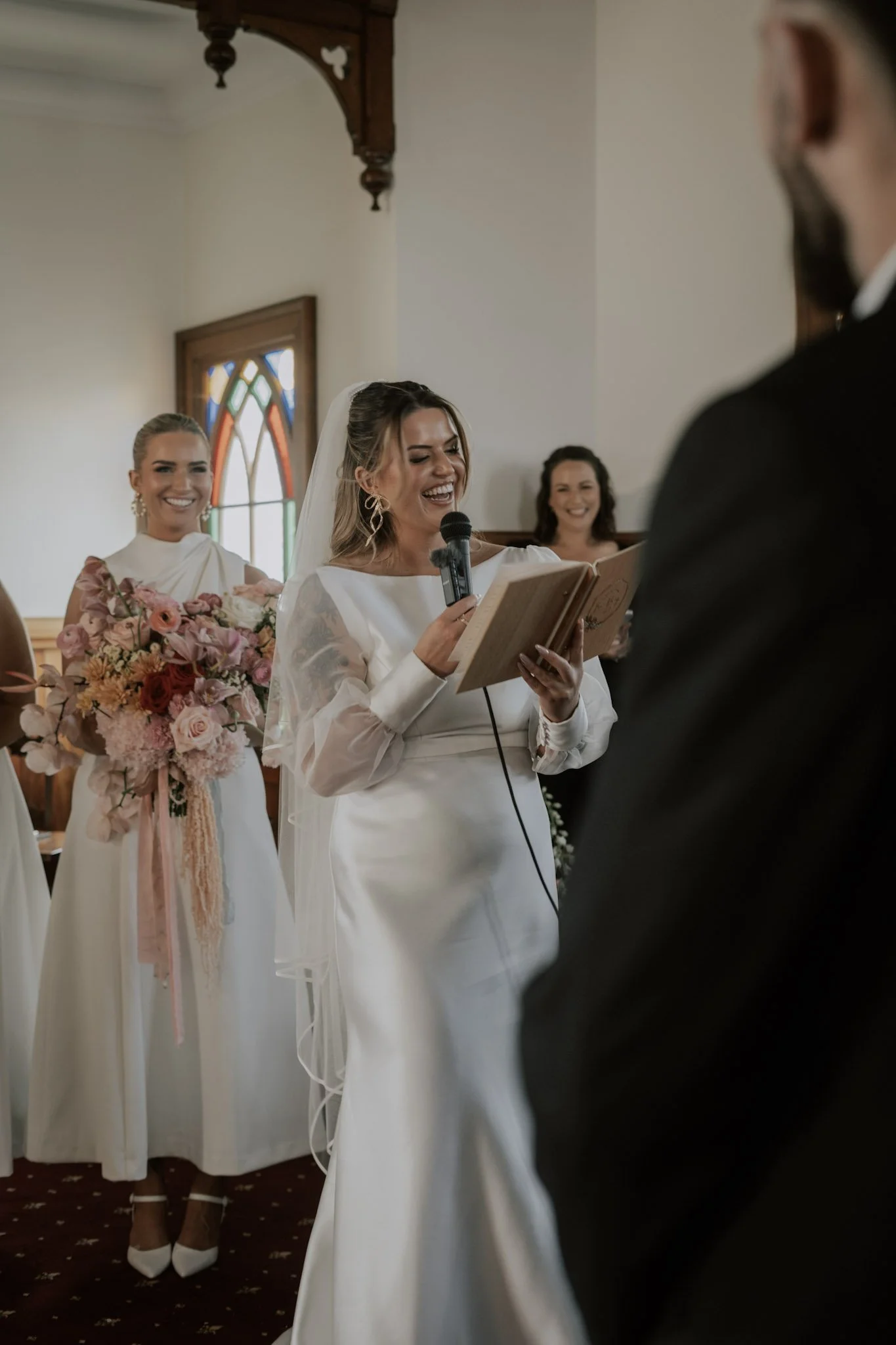 A bride giving a wedding vow reading inside a church, smiling and holding a microphone and a book, with bridesmaids holding bouquets in the background.