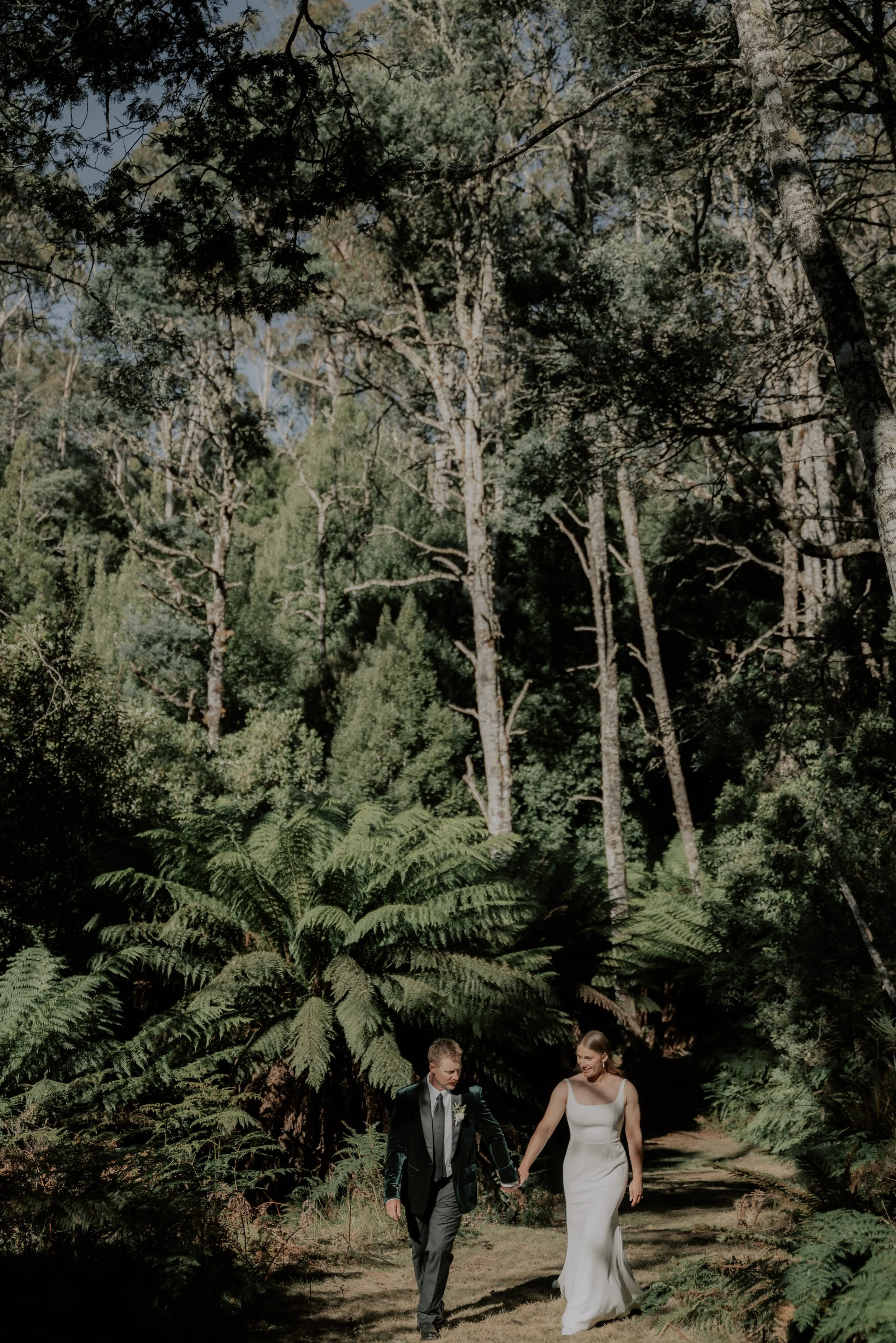 A newlywed couple walking hand in hand through a lush forested trail, surrounded by tall trees and ferns on a sunny day.