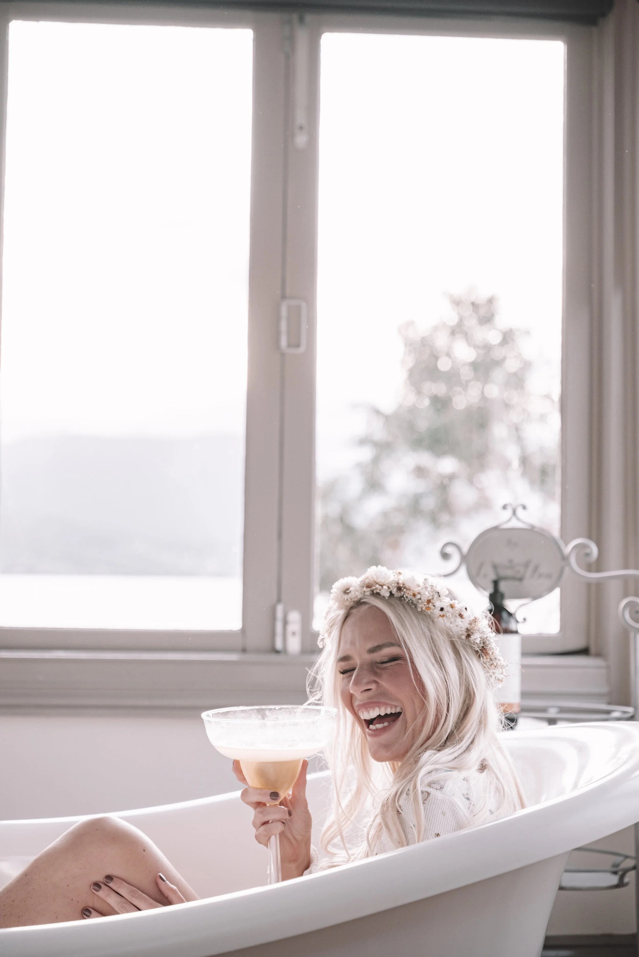 A woman with blonde hair, wearing a flower crown, is sitting in a bathtub, holding a champagne glass, and laughing.